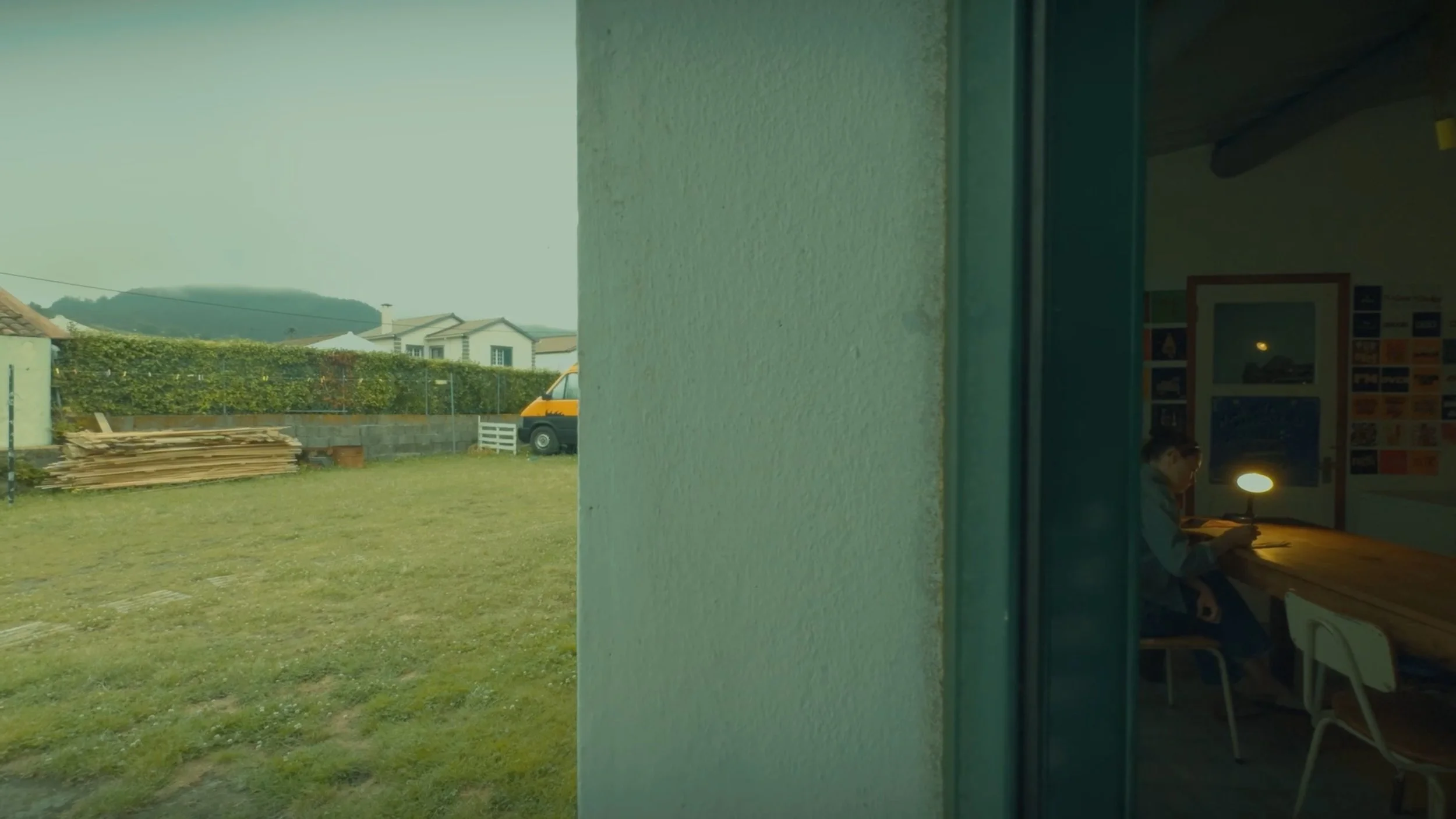 View through a window showing a grassy yard with wooden pallets, a yellow vehicle, and houses in the background under a cloudy sky. Inside, a person is sitting at a desk with a lamp, reading a book.