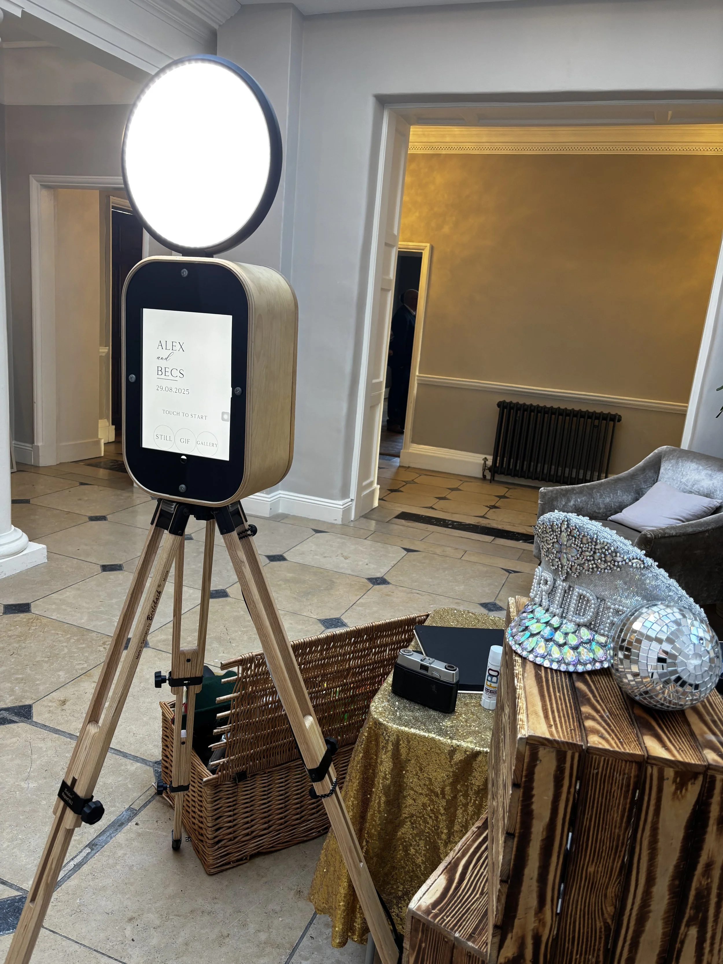 Photo of a photo booth setup with a ring light, tablet screen, and a wedding-themed prop on a wooden table, at Rockbeare Manor.