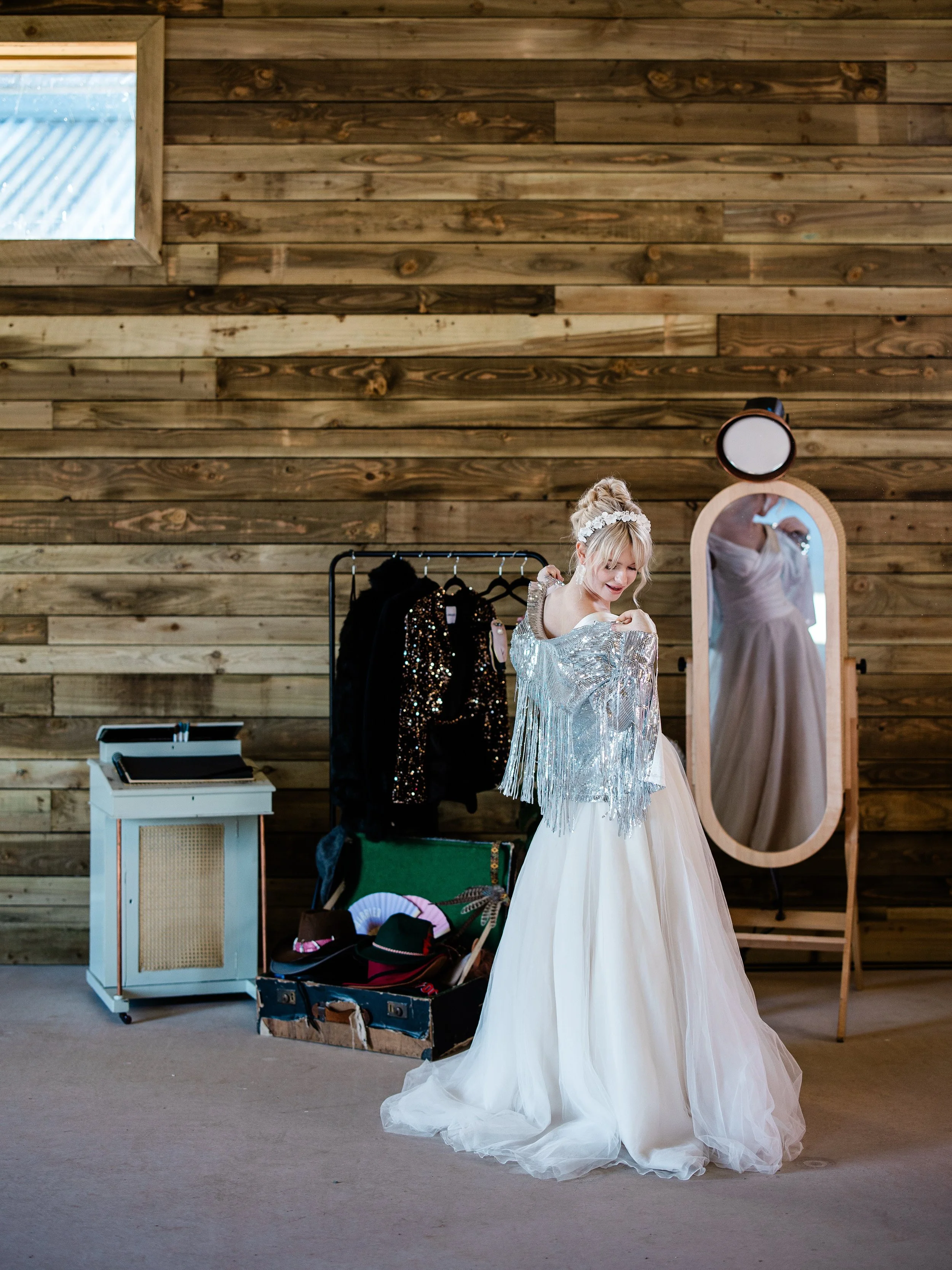 A woman in a wedding dress trying on a silver, fringed shawl in front of a rustic photo booth with vintage props.