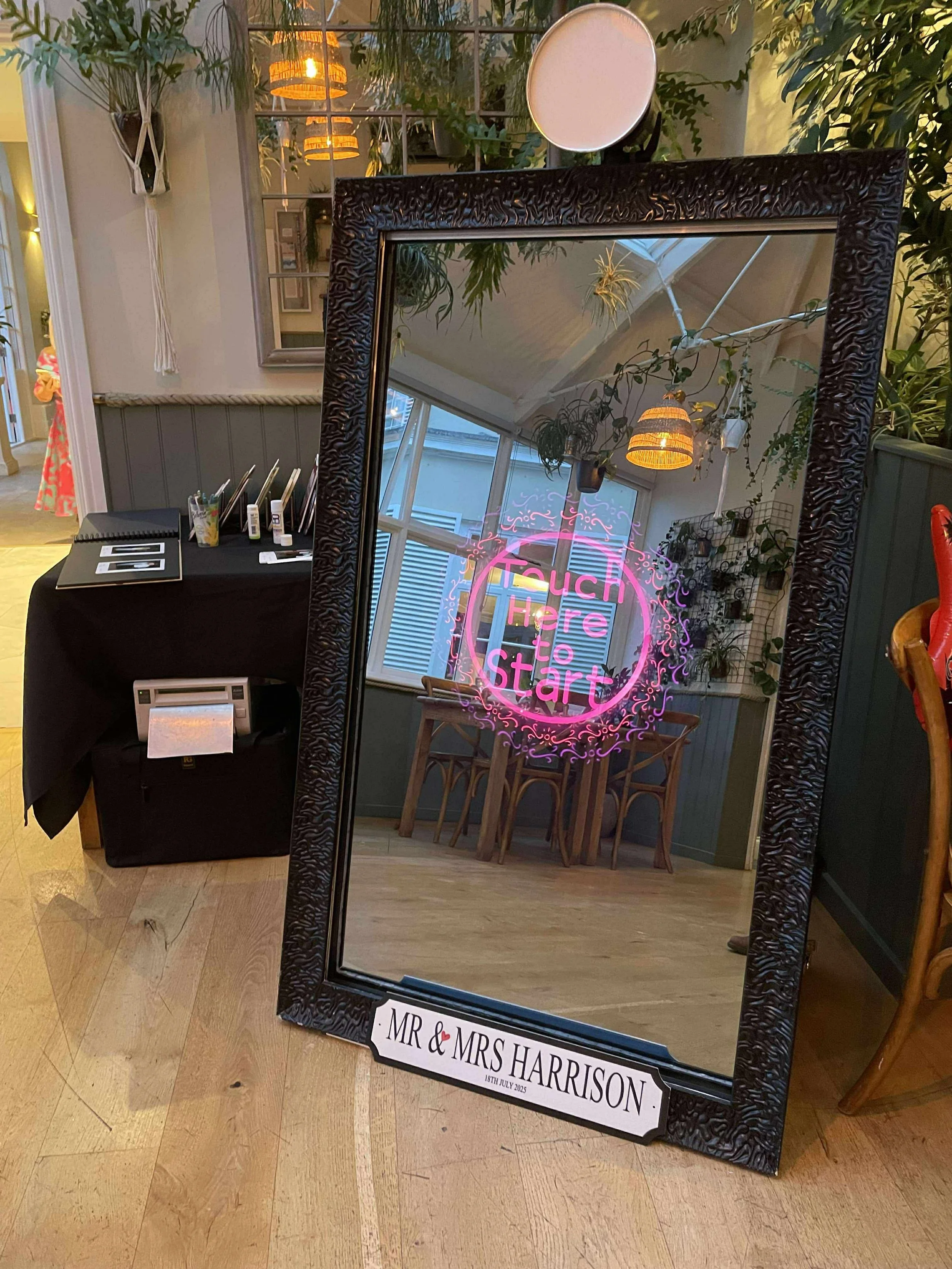 A magic mirror photo booth with a black textured frame, featuring an interactive screen that says "Touch Here to Start" reflected in it.
