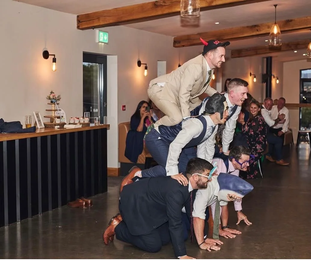 Group of people forming a human pyramid at a celebration, in front of a magic mirror photo booth with some wearing costumes and props.