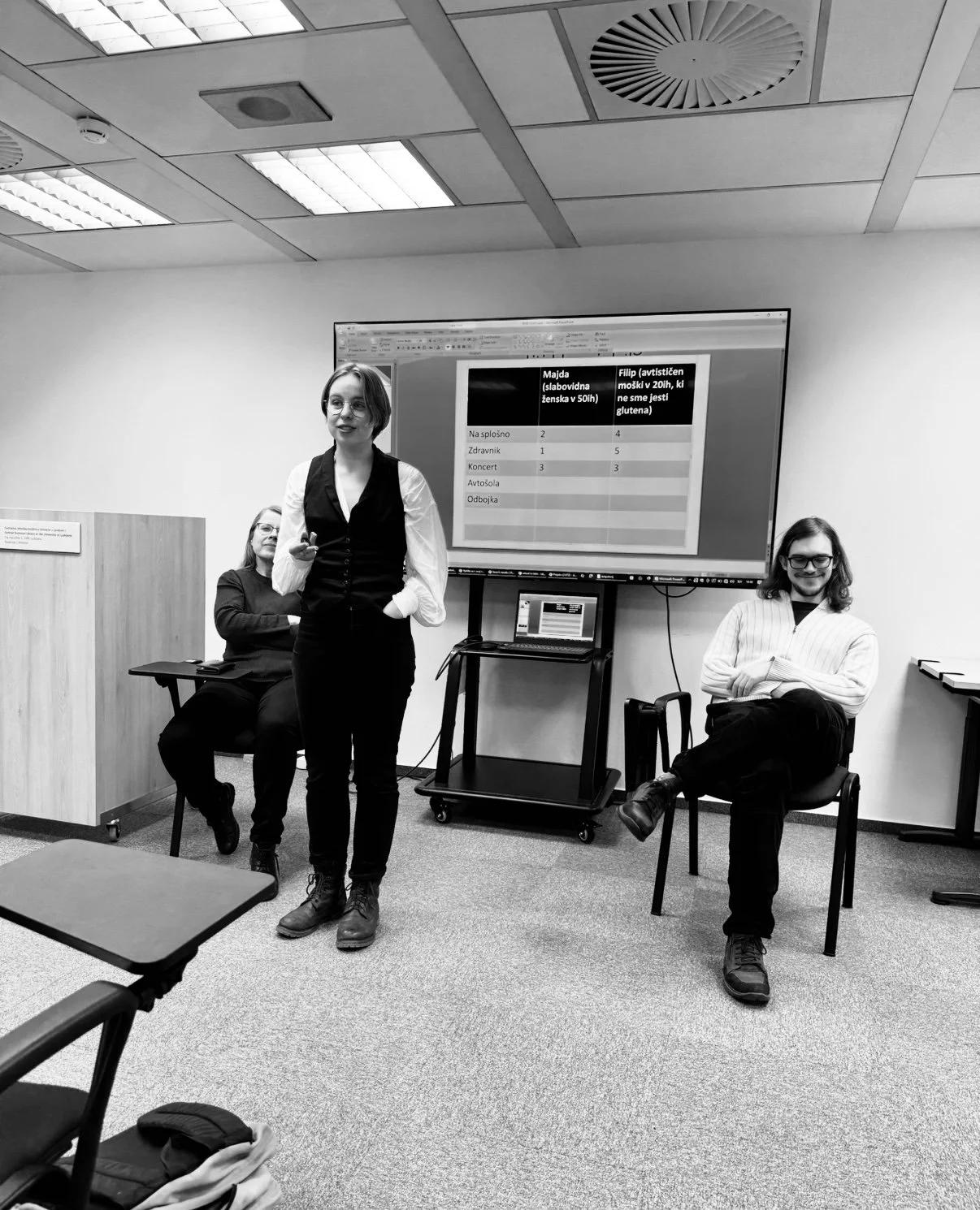 A woman giving a presentation in a conference room, with two seated women listening. A large screen displays a table with Slovene text and numbered columns. The room has ceiling lights and a minimalist office setup.