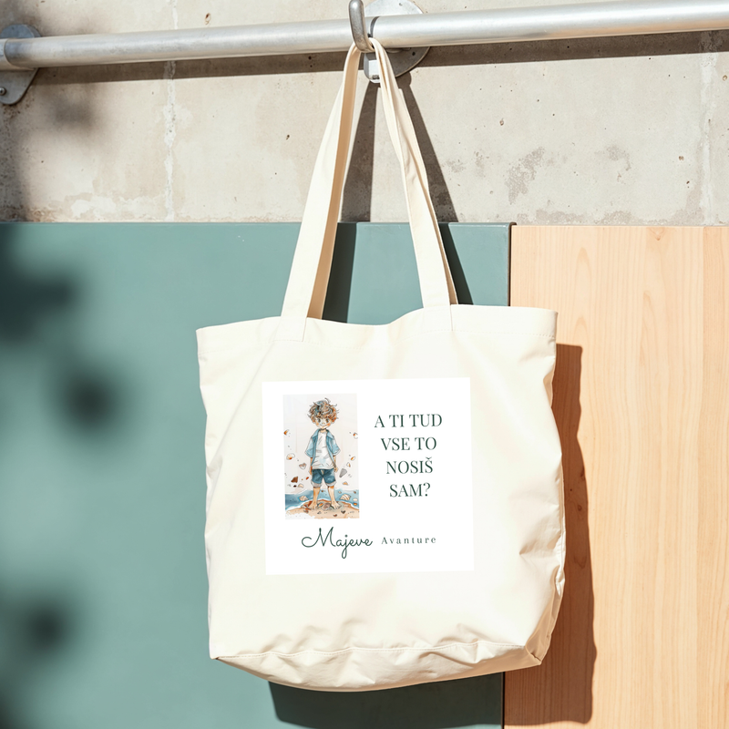 A white tote bag hanging on a metal rail against a wall with a green and wooden panel. The bag has a printed illustration of a person with gray hair and the text in a foreign language, possibly Slovenian, with the words 'Majeve Avanture'.