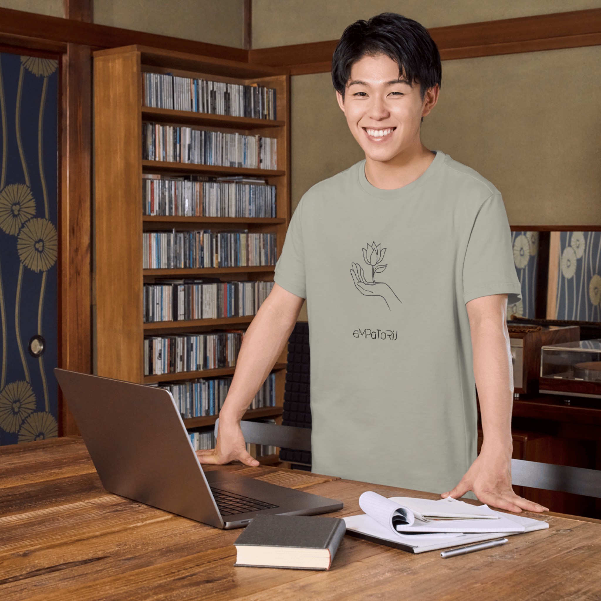 A young man smiling, standing behind a wooden desk with a laptop, notebook, and papers, in a room with a bookshelf filled with CDs and a decorative wall.