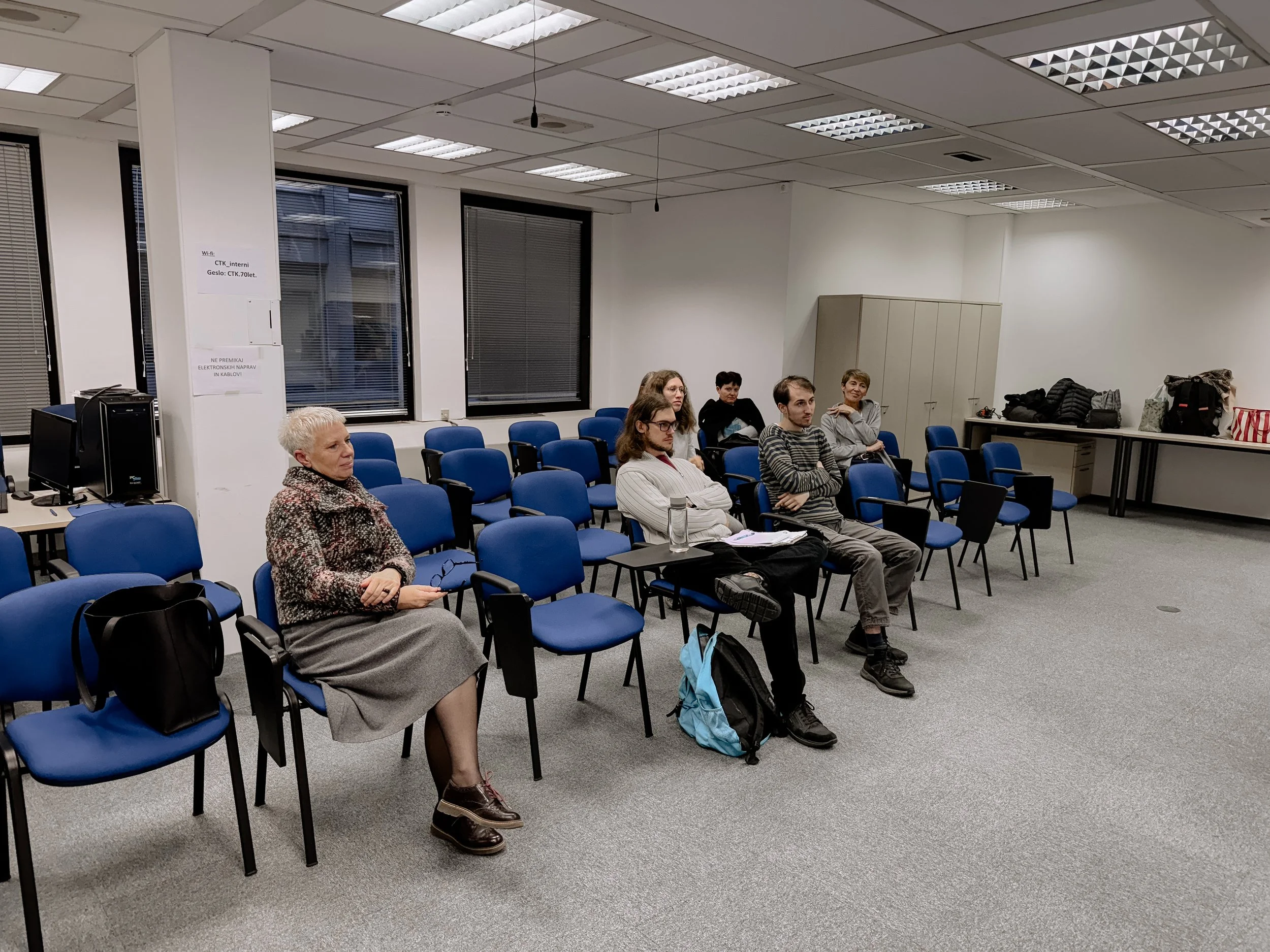 A classroom or conference room with blue chairs arranged in rows, some occupied by people. A woman in a gray skirt and patterned sweater sits on a chair, while five young adults sit nearby, some with backpacks and notebooks. The room has white walls, large windows with blinds, and gray carpeting. To the side, there are tables with bags and jackets, and a computer setup near the window.