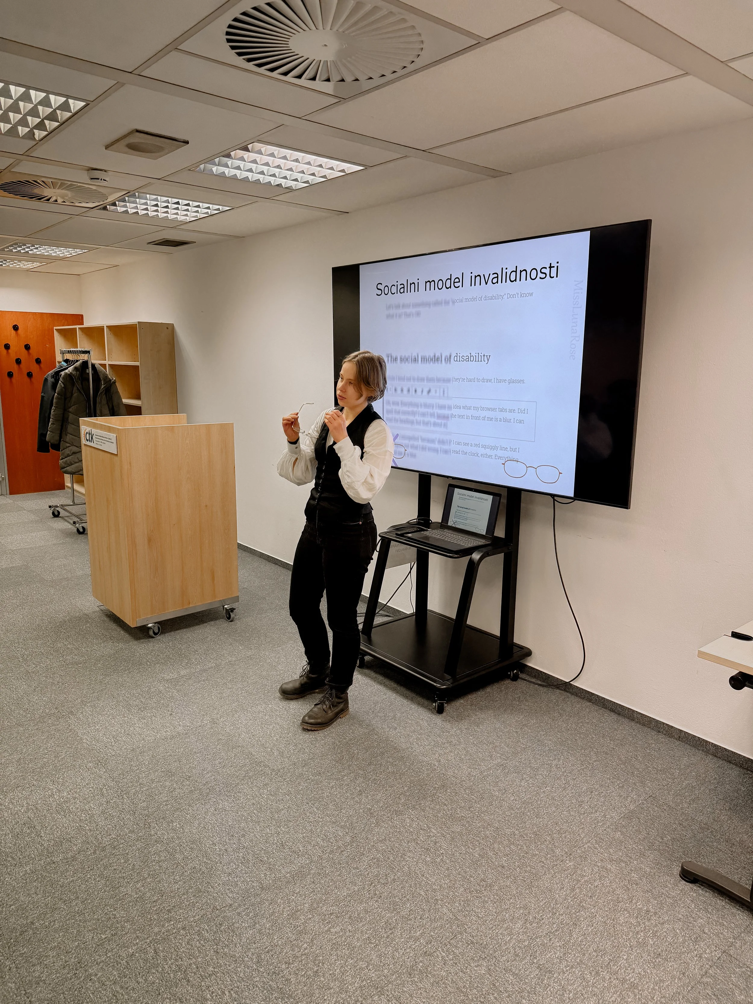 A woman giving a presentation in a conference room with a large screen displaying a slide titled 'Socialni model invalidnosti'. She is standing in front of the screen, holding glasses, with a laptop behind her on a stand. Behind her, a wooden coat rack with jackets and a wooden storage unit are visible.