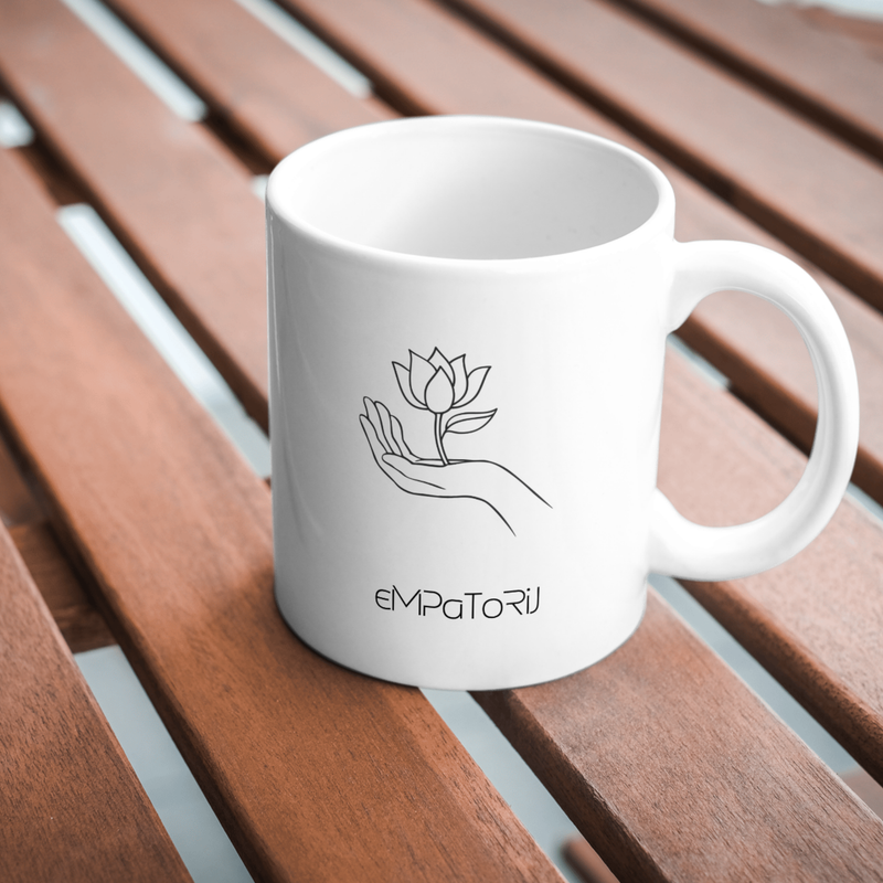 A white ceramic mug with a minimalist line drawing of a hand holding a blooming lotus flower. The mug is placed on a wooden slatted surface.