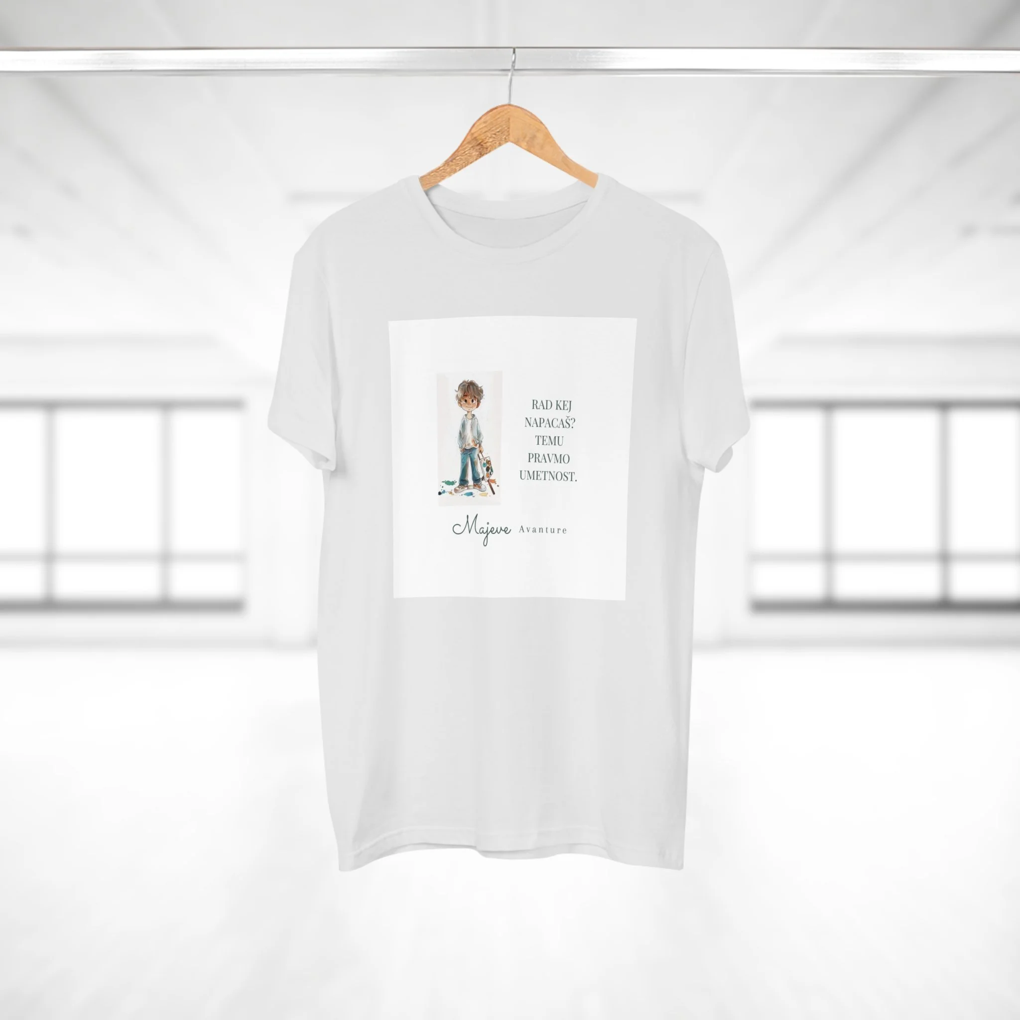 White T-shirt with a printed illustration of a young boy and text in a different language hanging on a wooden hanger in a bright, modern room with large windows.