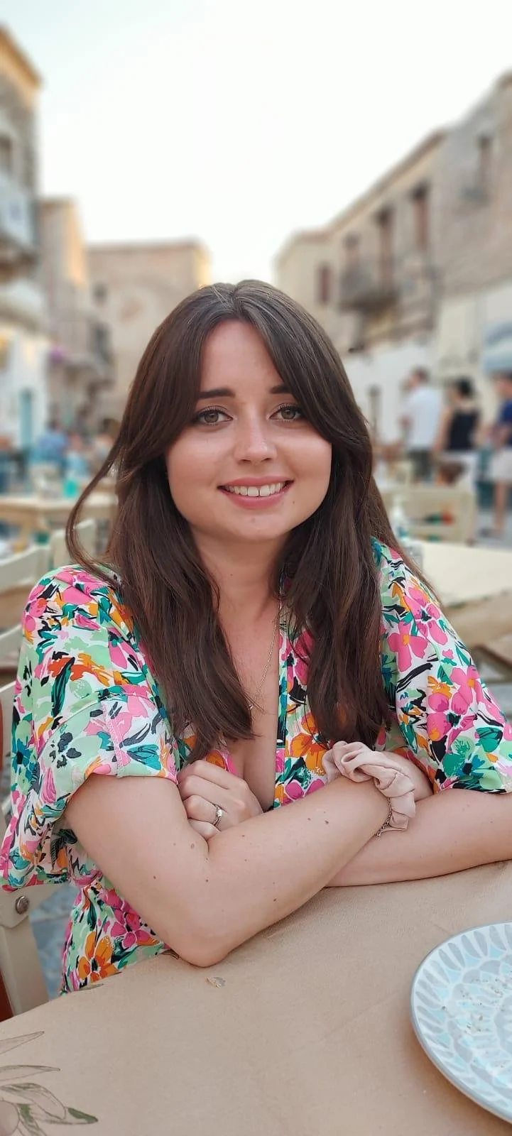 A young woman with long brown hair, smiling, sitting outdoors at a table in a lively area