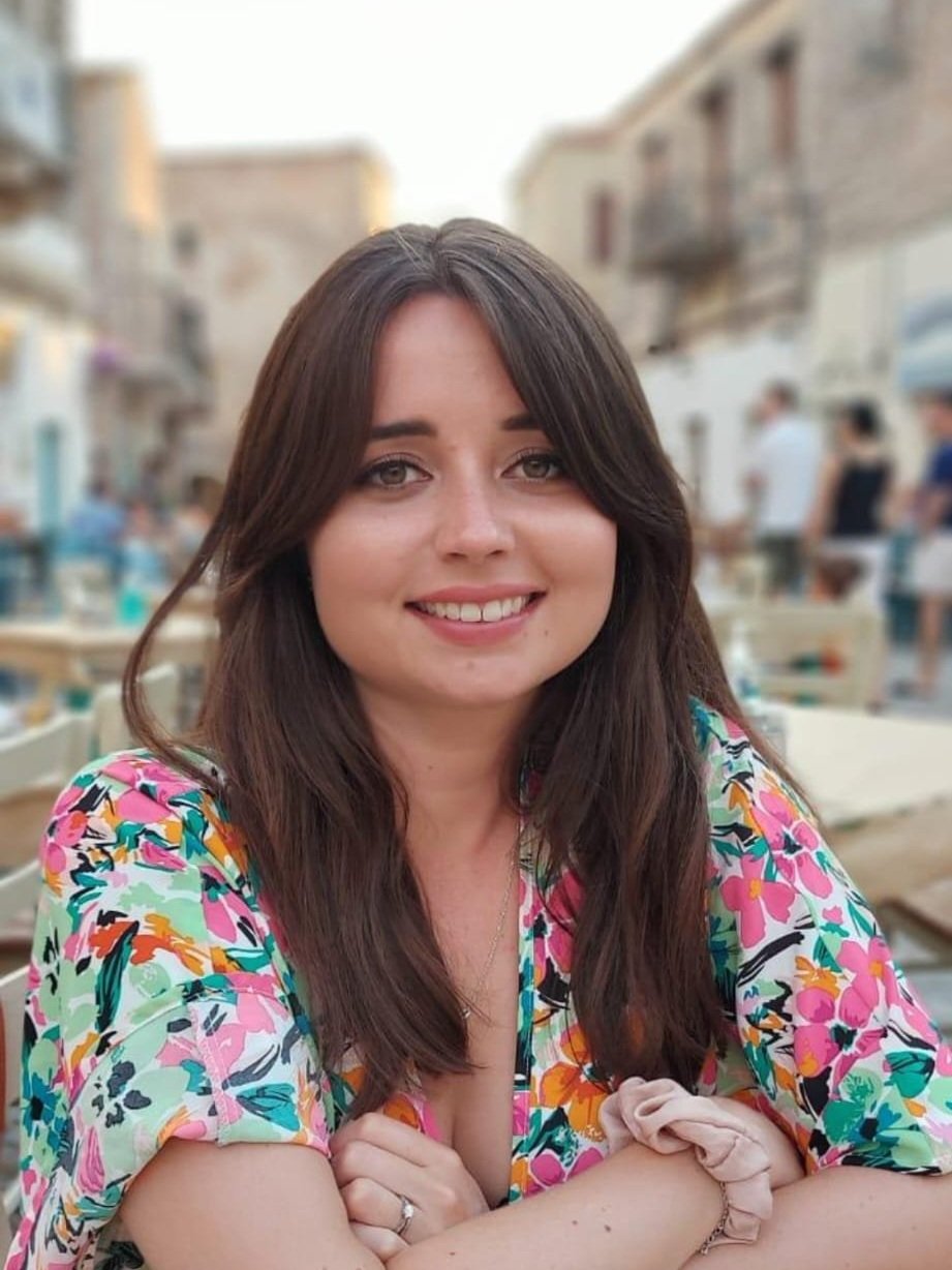 A young woman with long brown hair smiling, sitting outdoors at a cafe during daytime, wearing a colorful floral dress.