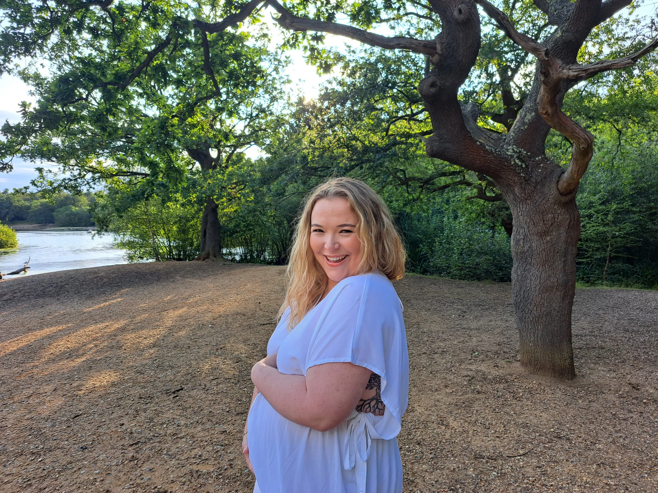 A smiling woman with blonde curly hair wearing a white dress standing outdoors near a river and trees during daytime.