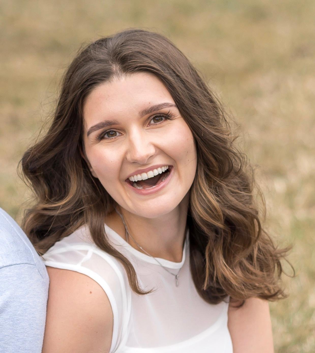 A young woman with long, wavy brown hair smiling outdoors.