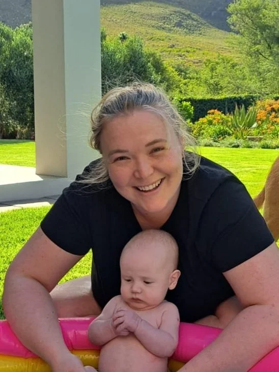 A woman smiling outdoors with a baby in a small pool, green grass, trees, and a hillside in the background.