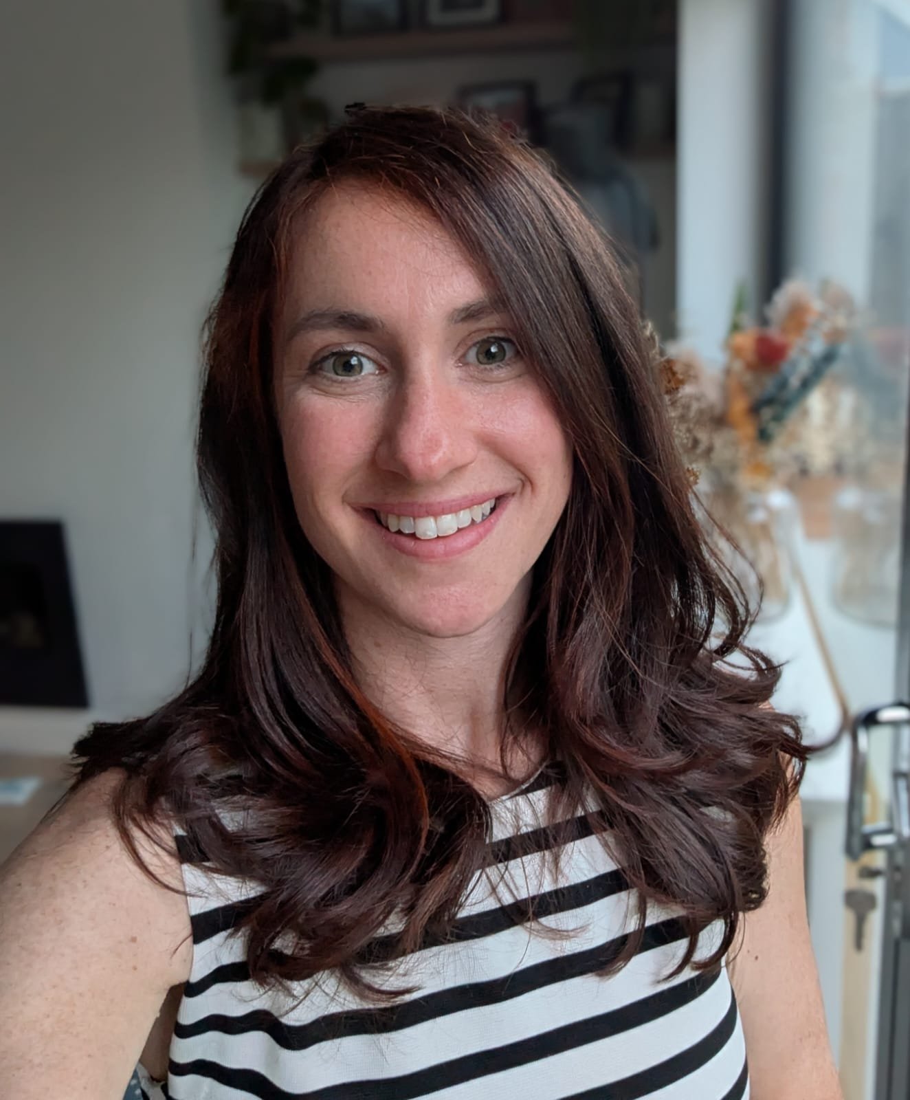 A smiling woman with long wavy brown hair wearing a black and white striped sleeveless top, standing in a well-lit room with a window and a blurred background of flowers and picture frames.