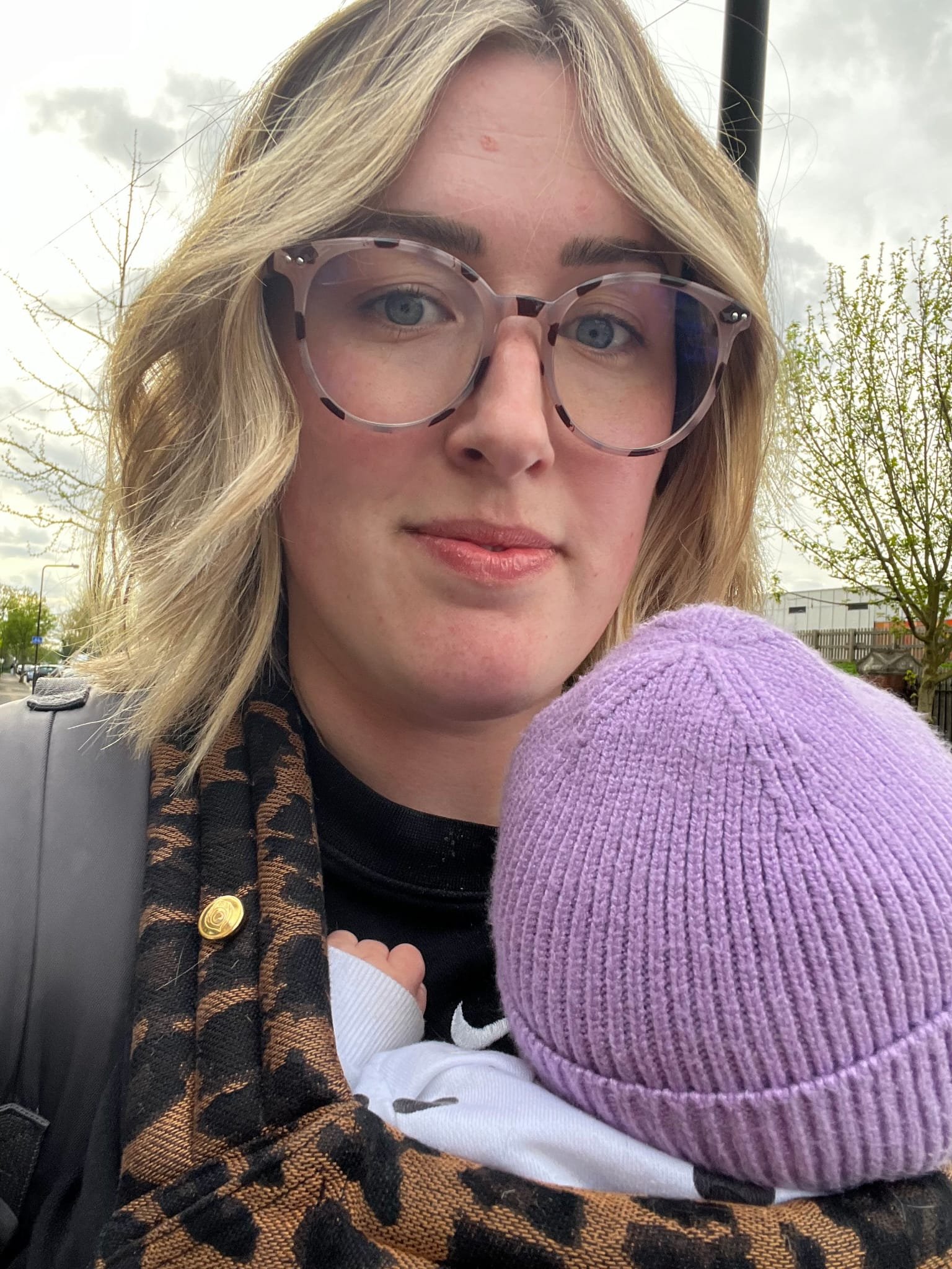 A young woman with blonde hair, wearing large patterned glasses, holding a baby in a purple knit hat; they are outdoors on a cloudy day.