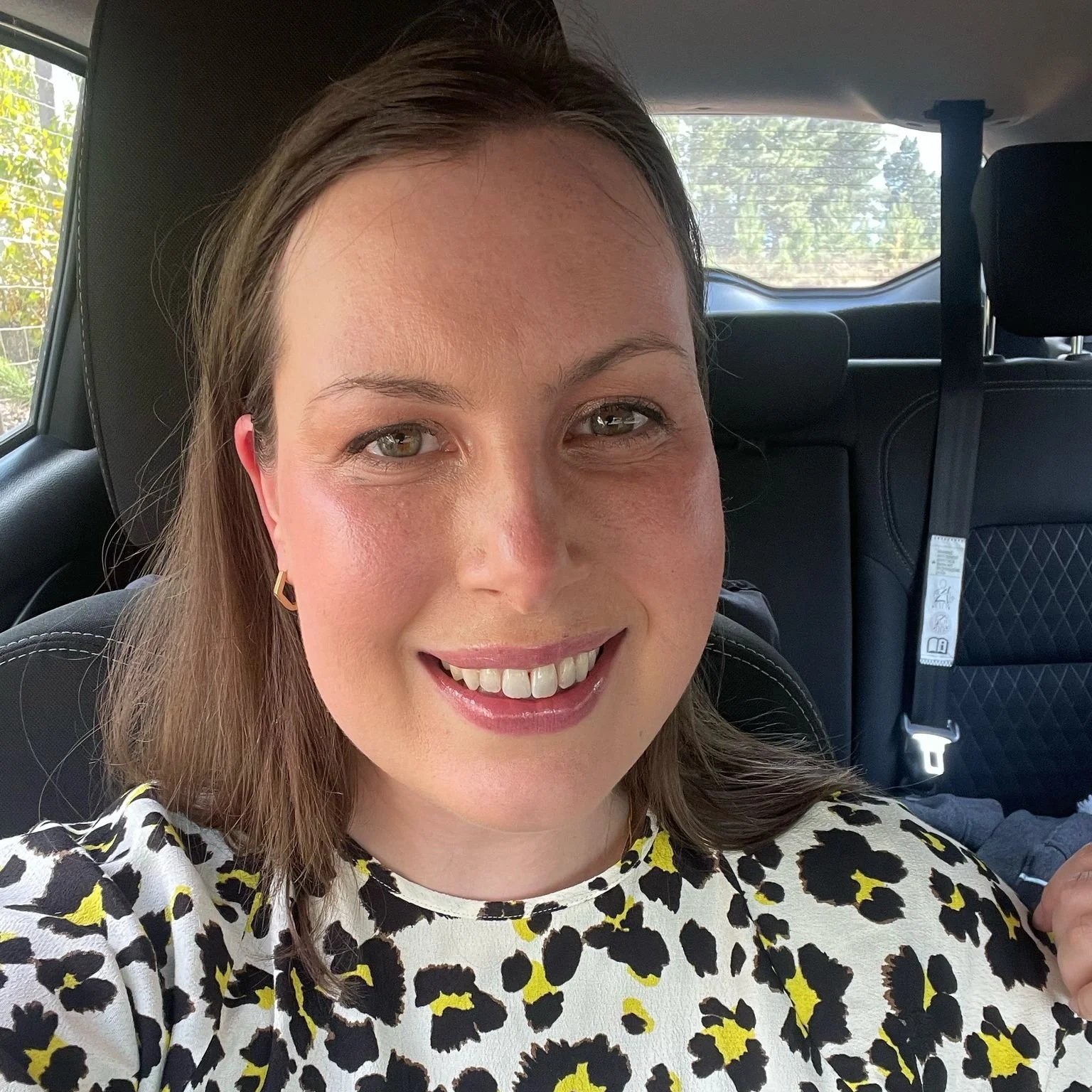 A woman with brown hair, wearing a leopard print top, smiling inside a vehicle.