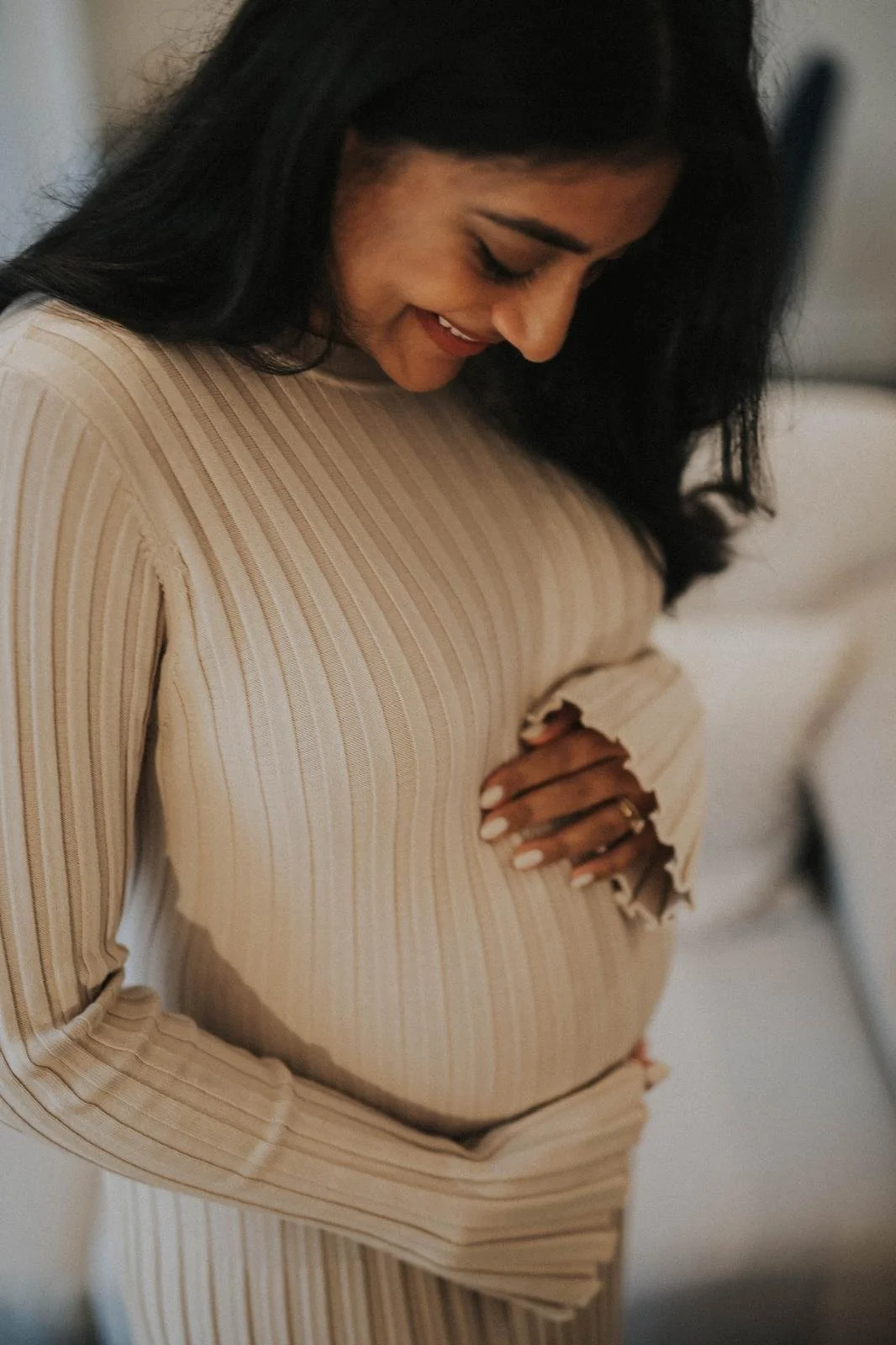 A pregnant woman with long dark hair smiling, wearing a beige striped long-sleeve top, gently touching her belly.