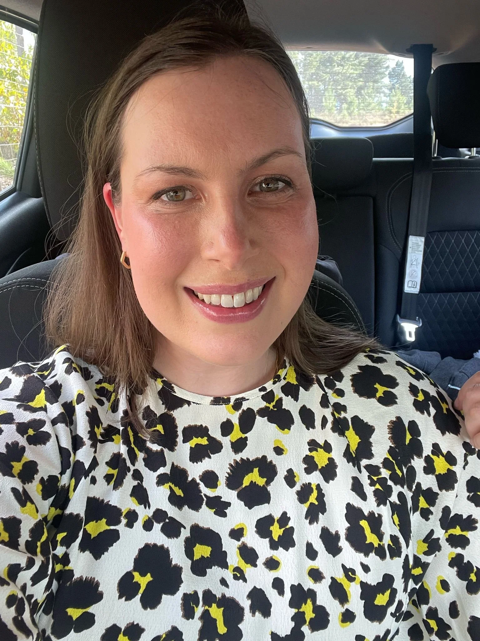 A woman smiling in a car, wearing a black and white leopard print top with yellow accents, and gold earrings.