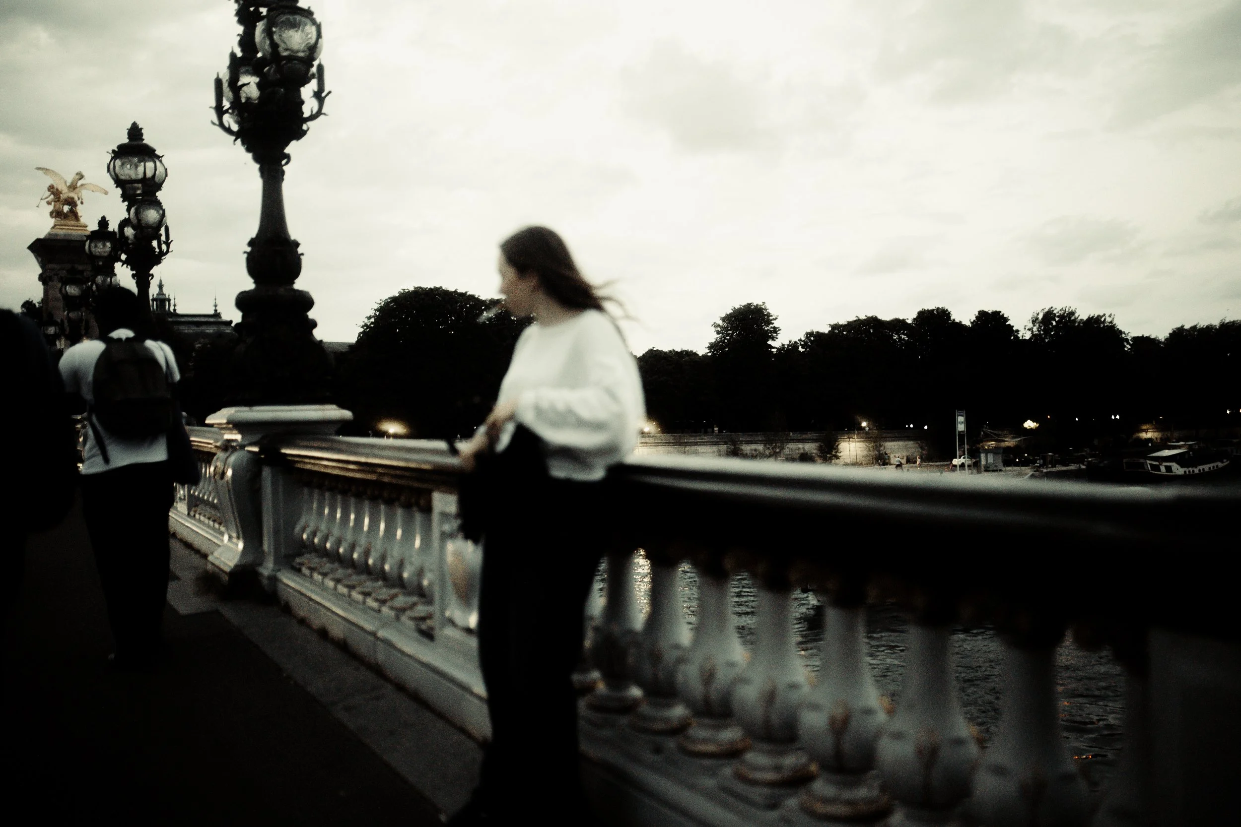 Una donna si appoggia alla ringhiera di un ponte affacciato su un fiume al tramonto, con diverse persone che passeggiano e lampioni decorativi lungo la balaustra.