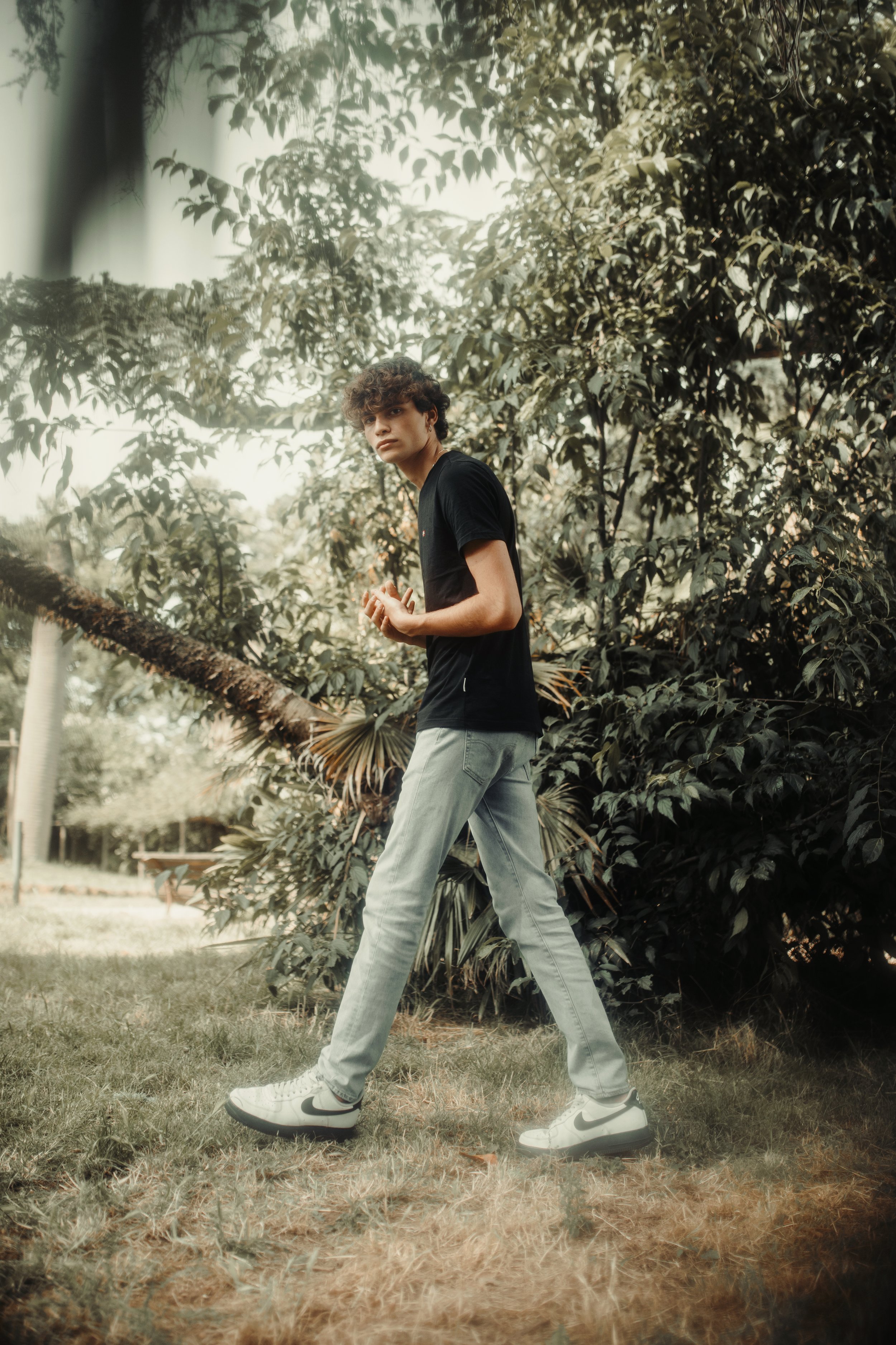 Uomo cammina in un parco, indossando jeans chiari e maglietta nera, con una foresta sullo sfondo.