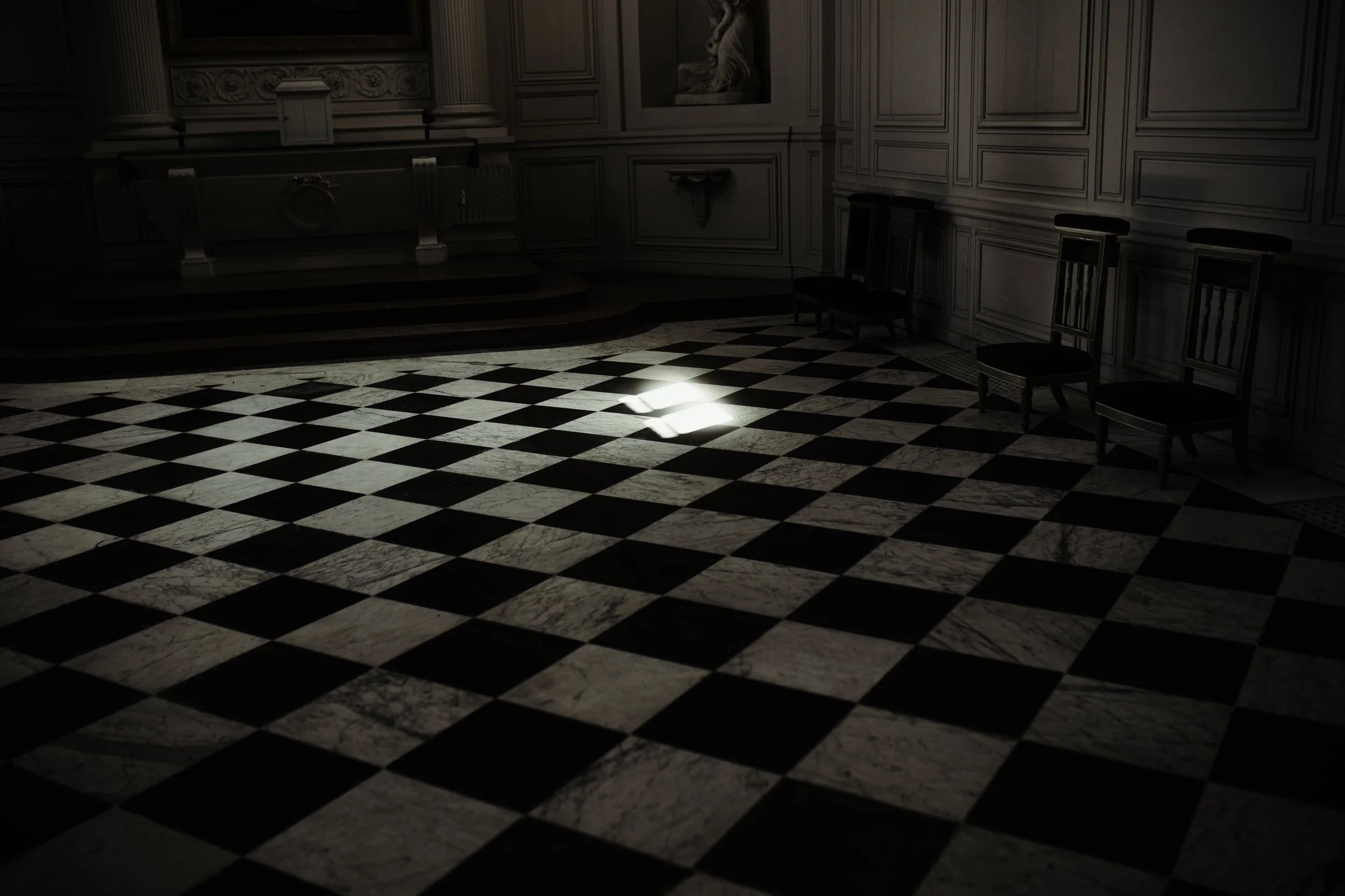 Stanza vuota con pavimento a scacchi in marmo bianco e nero, tre sedie e un'area leggermente illuminata su pavimento.