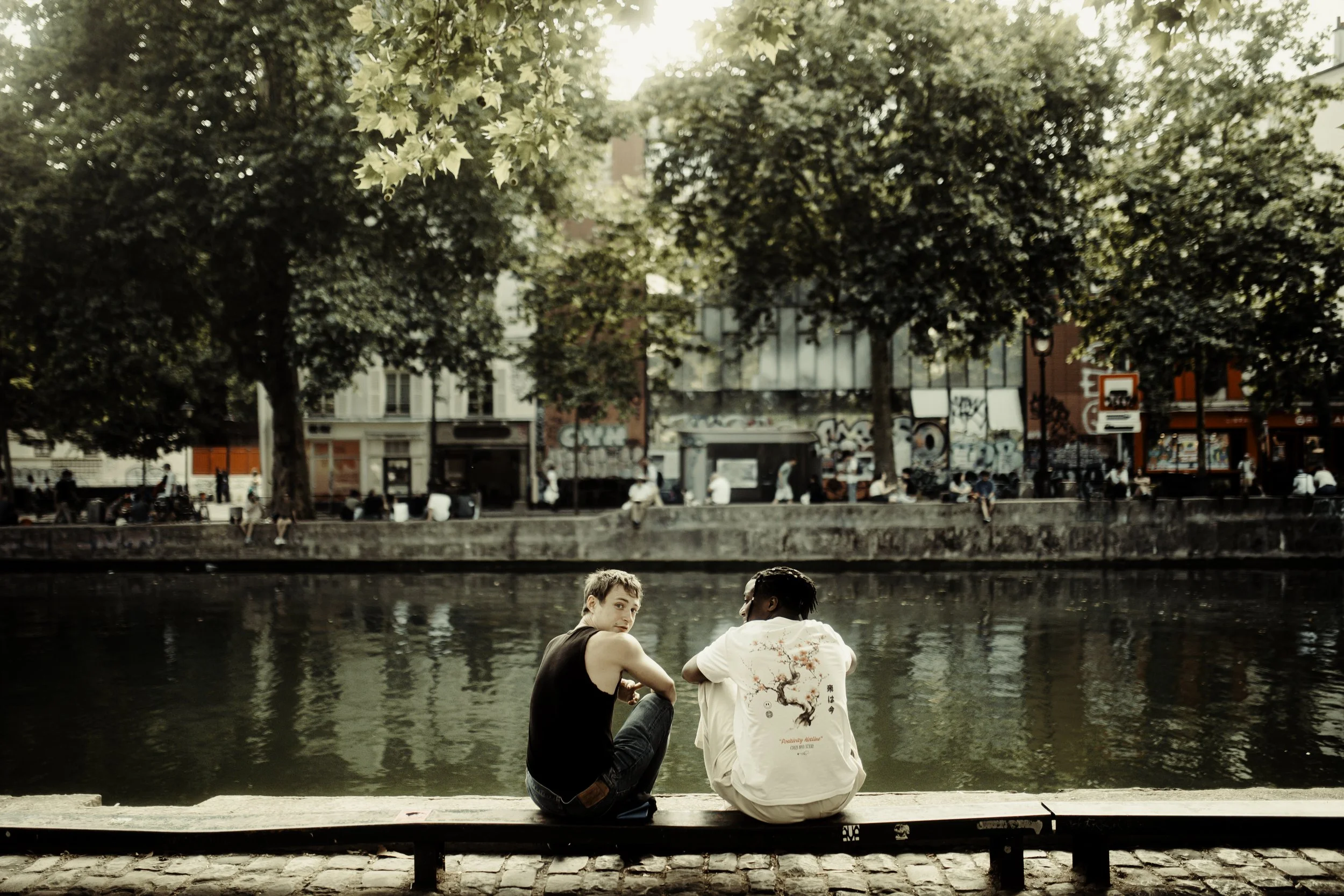 Due giovani seduti su una panchina accanto a un fiume, in una zona urbana con alberi e edifici sullo sfondo.