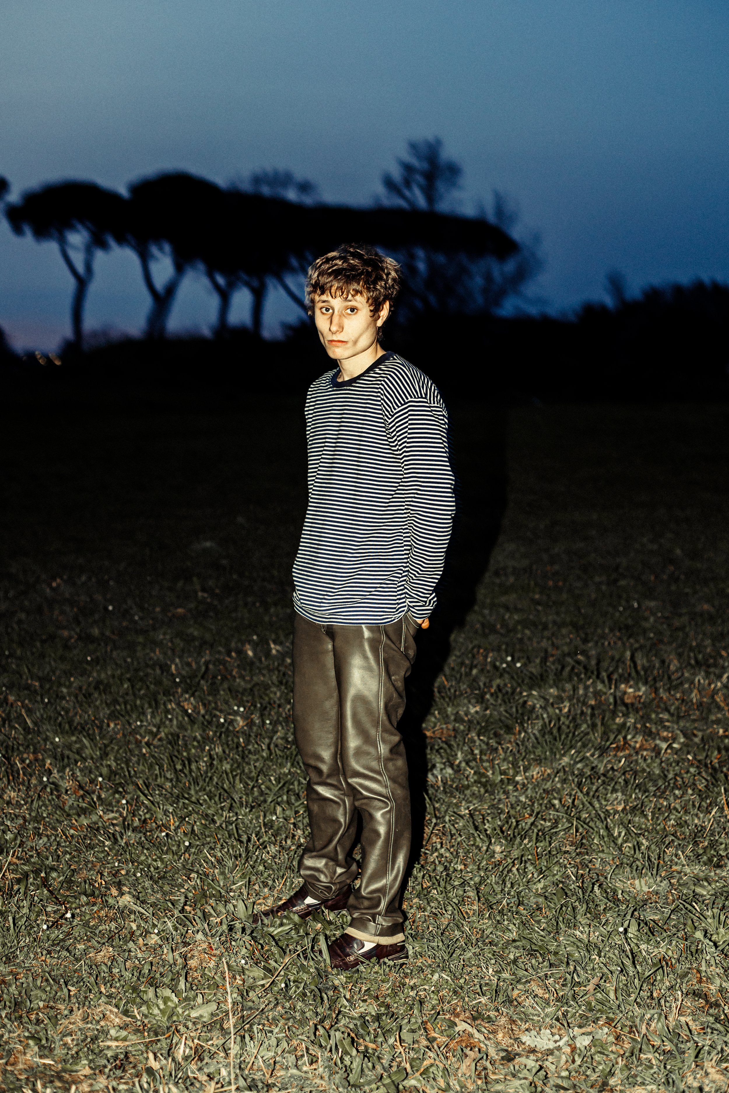 Persona in campo con maglia a righe, pantaloni lunghi e alberi sullo sfondo.