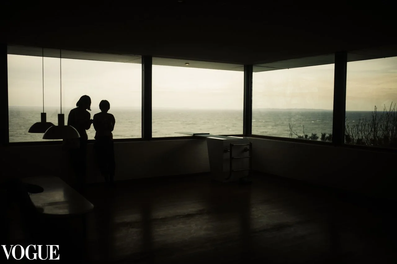 Due persone in silenzio guardano fuori da grandi finestroni una vista sull'oceano, in un ambiente interno oscurato, con siluette contro una luce naturale