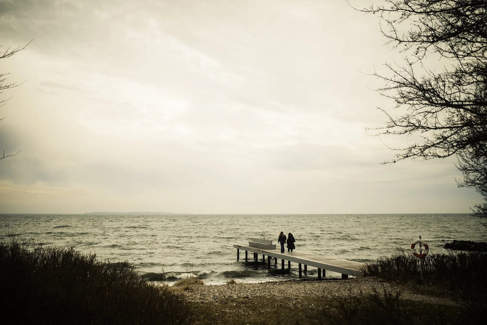 Due persone camminano su un molo di legno verso il mare sotto un cielo nuvoloso, con alberi spogli ai lati.