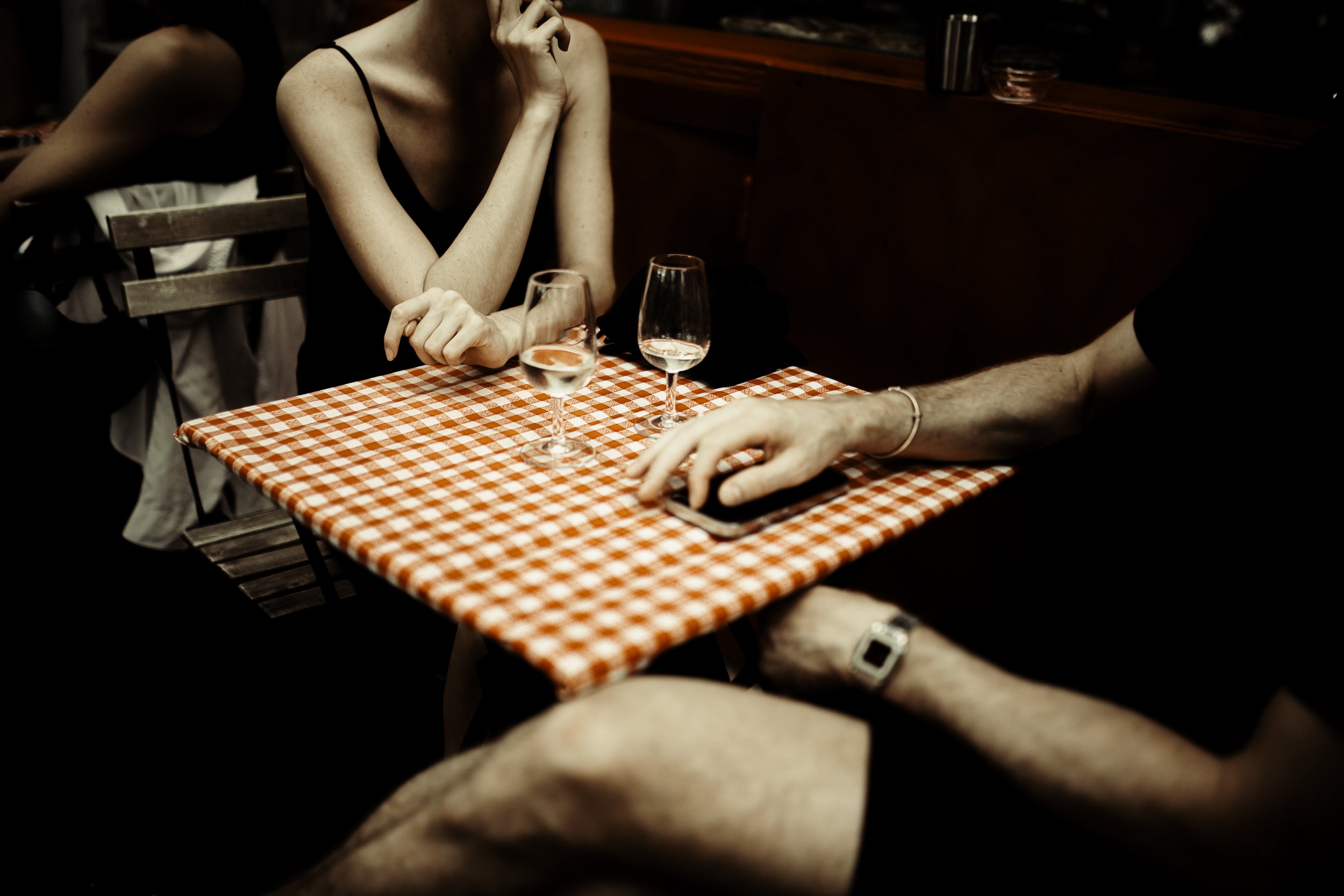 Due persone sedute a un tavolo da ristorante con tovaglia a scacchi rossi e bianchi, uno con un braccialetto e l'altro con un orologio, ci sono due bicchieri con acqua sulla tavola.