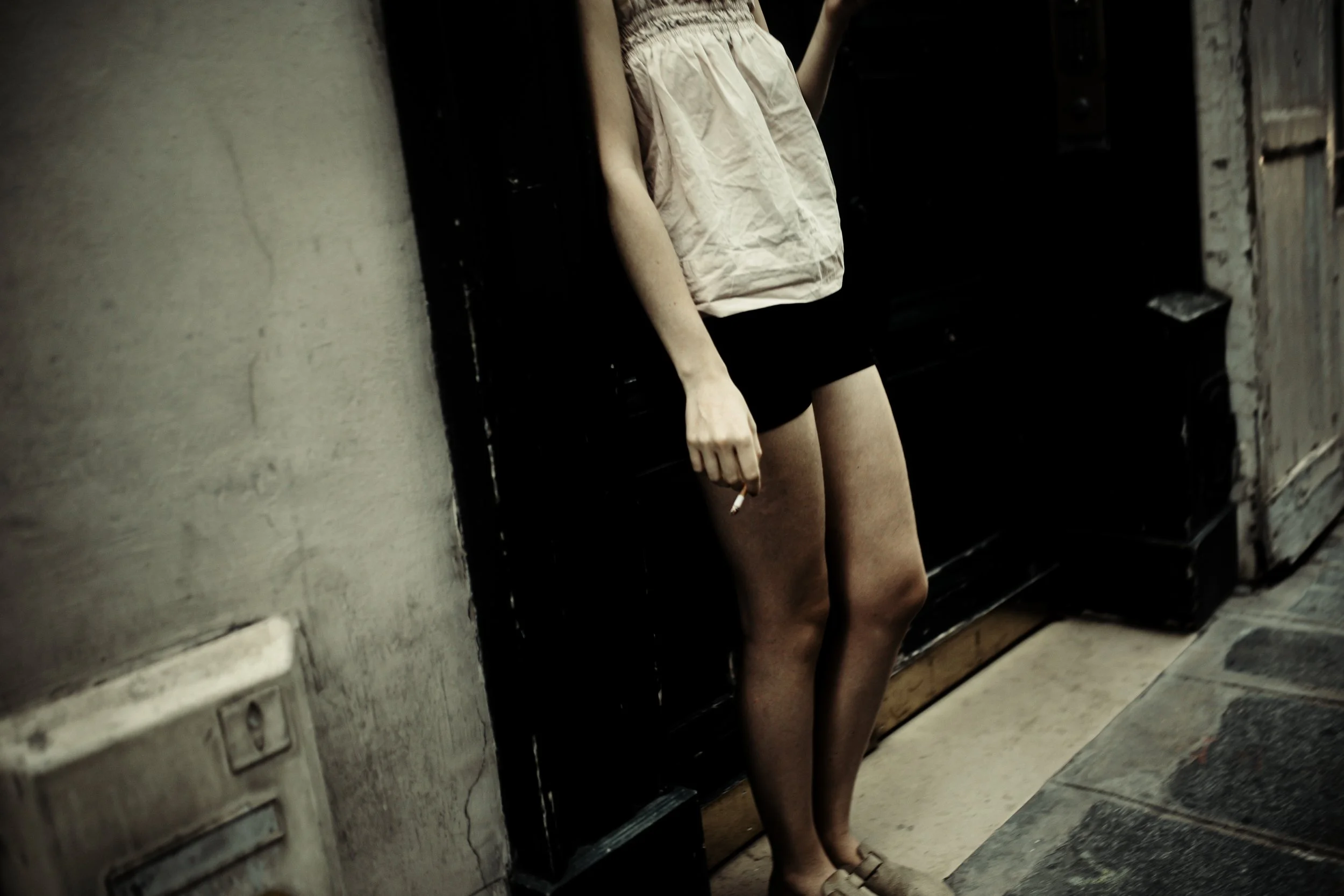 Una donna indossa un abbigliamento casual con una maglietta beige, pantaloncini neri e scarpe beige, mentre si trova in un'ambientazione urbana, appoggiata con la schiena a una parete scura e un tubo nero di fianco.