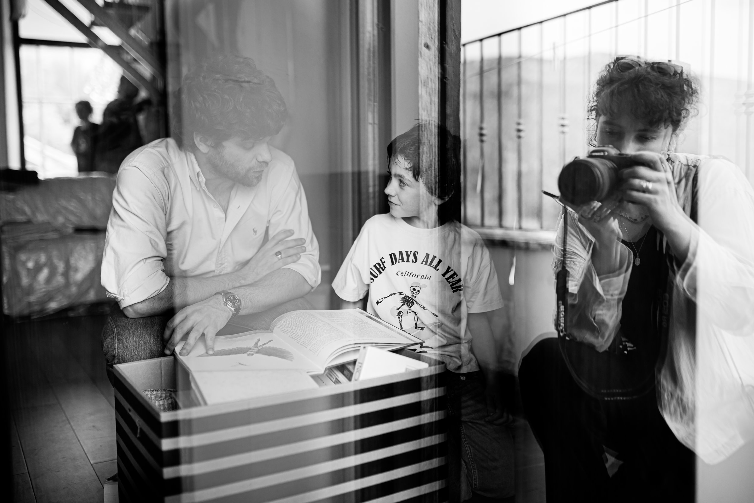 Scena in bianco e nero di tre persone dietro un vetro; un uomo e un bambino sono seduti e guardano un libro aperto, mentre una donna fotografa la scena.