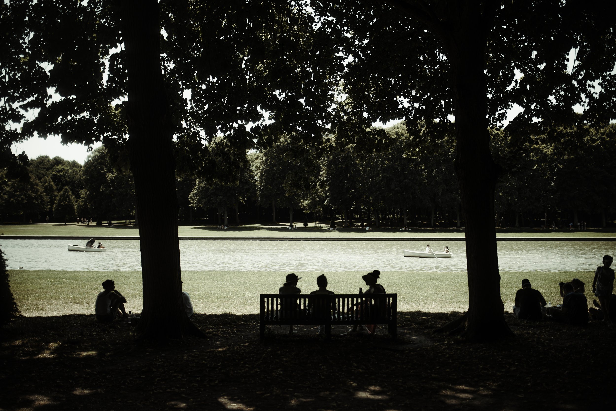 Persone che si rilassano e conversano sedute su una panchina sotto gli alberi vicino a un lago con barche a remi, in un parco durante il giorno.