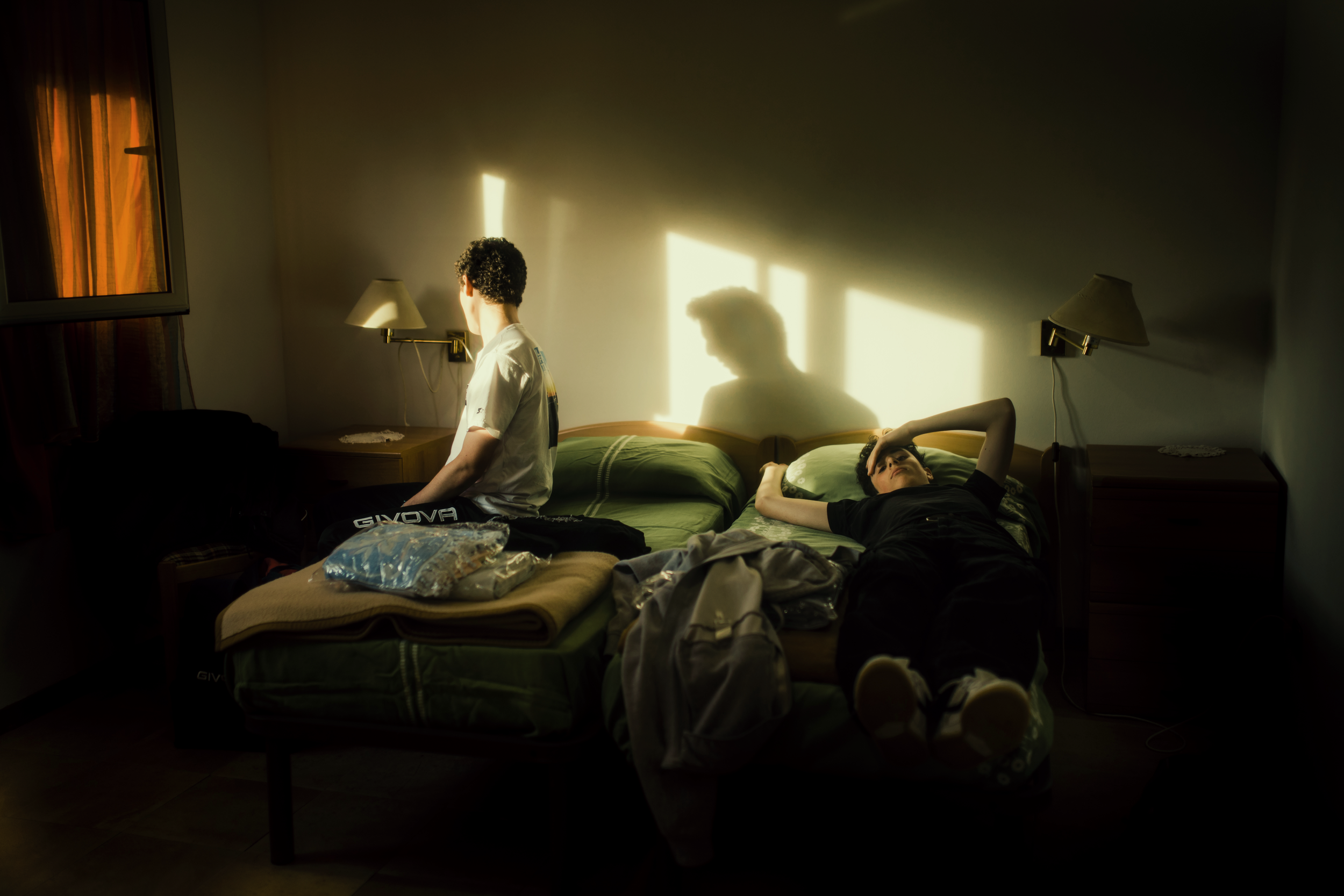 Una stanza d'albergo con due giovani, uno seduto sulla sponda del letto e l'altro sdraiato, con la luce del tramonto che crea un'ombra sulla parete.