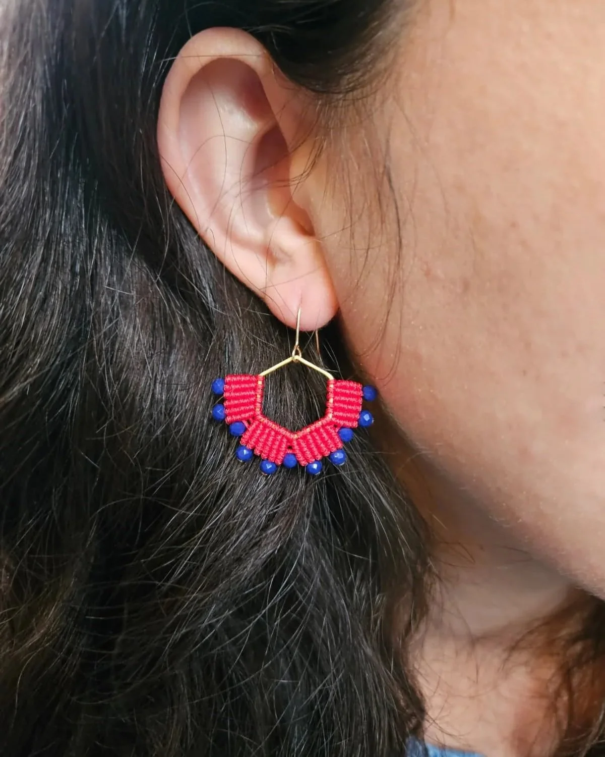A woman is wearing a large, colorful earring with a gold hoop, red woven material in a semi-circle shape, and small blue beads hanging from it.