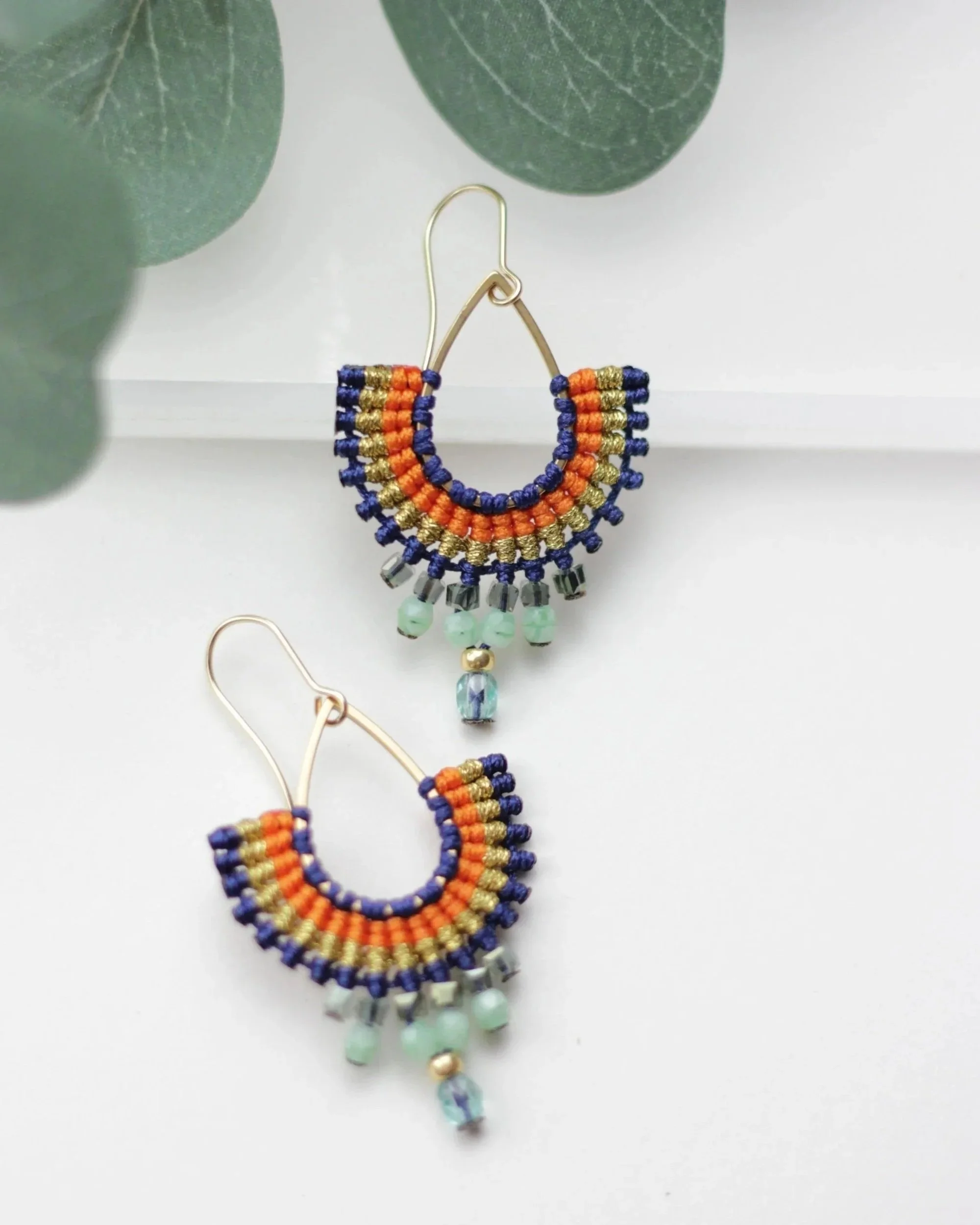 Colorful beaded earrings with orange, blue, gold, and green beads, featuring dangling beads at the bottom, placed on a white surface with green leaves in the background.