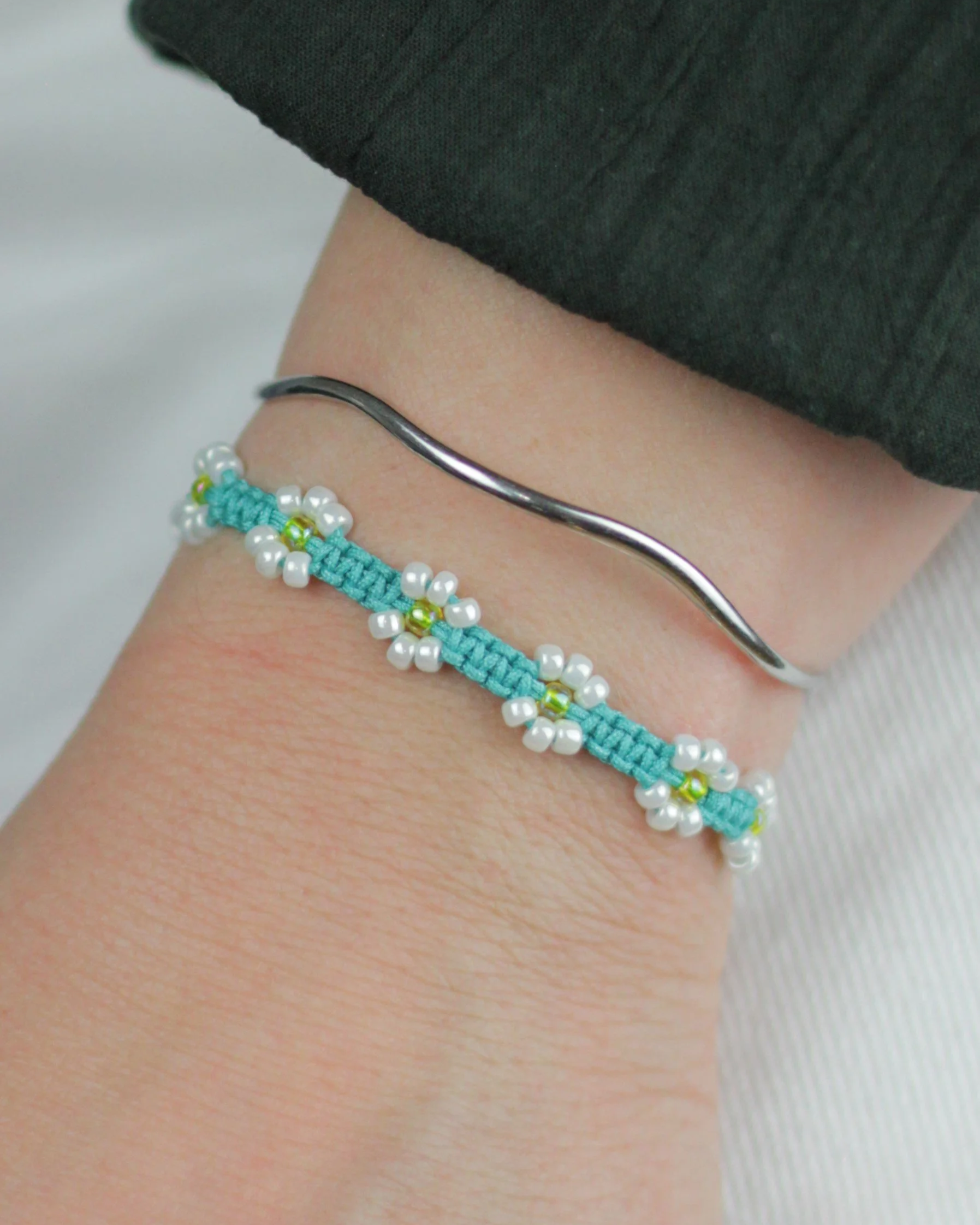 Two bracelets on a person's wrist: one is a simple, thin, silver band; the other is a turquoise woven band with small white and yellow beads forming floral patterns.