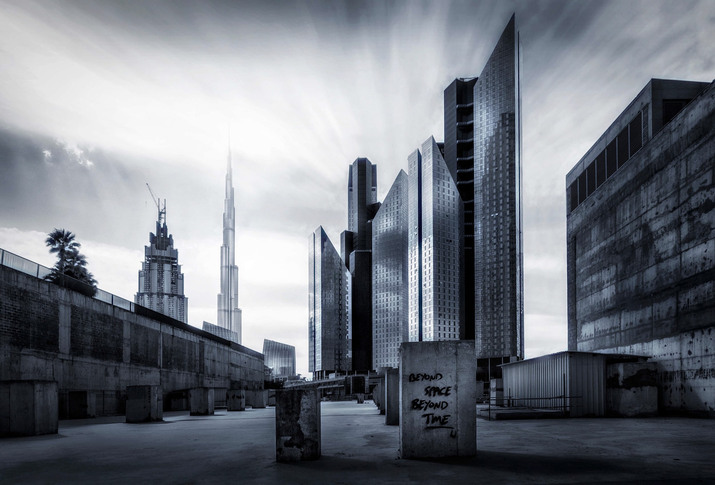 Black and white photograph of a cityscape with tall modern buildings, including the Burj Khalifa, under a cloudy sky. In the foreground, there is an open space with concrete structures and a sign that reads 'BEYOND SPACE BEYOND TIME.'