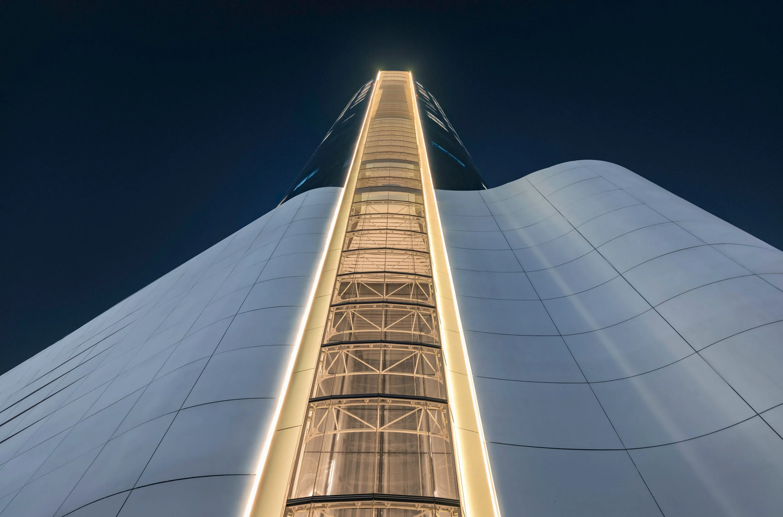 Looking up the lit glazed slot in the podium of Ciel Dubai Marina at night, the white cladding curving away on both sides.