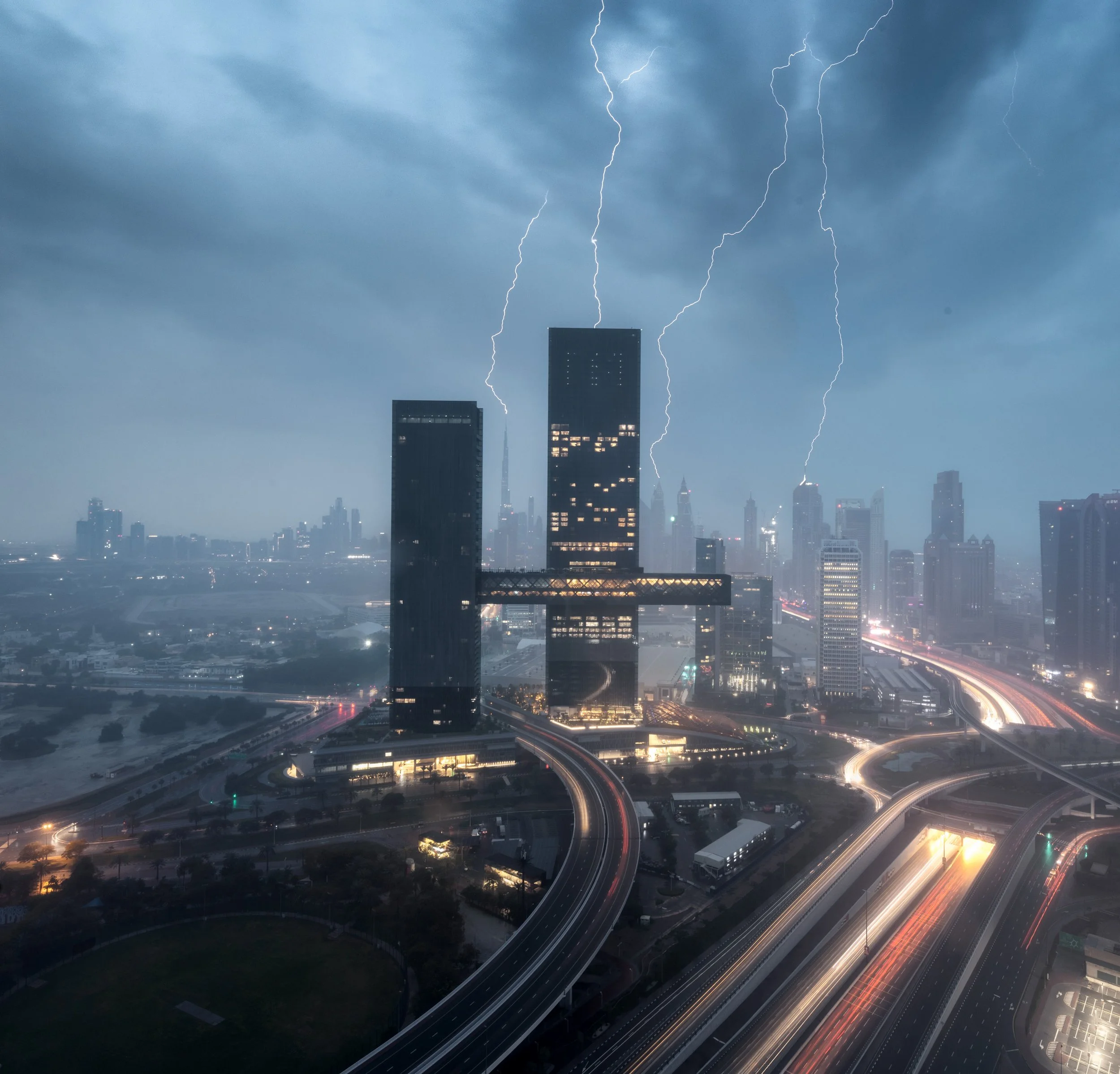 Nighttime cityscape with tall buildings and skyscrapers, lightning strikes in the stormy sky, highway with moving cars creating light trails, and illuminated streets.