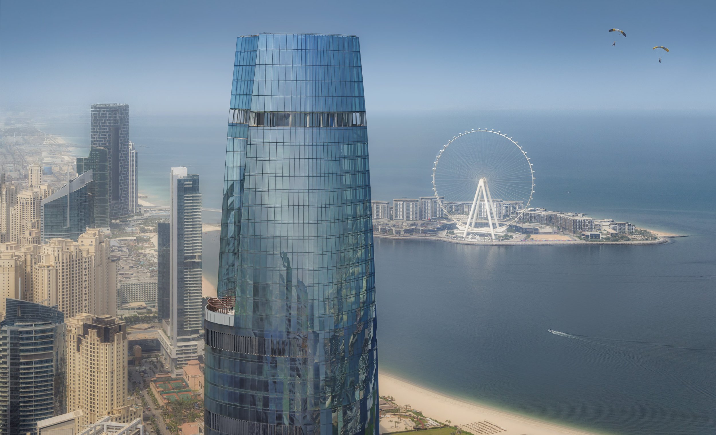 A city skyline with tall modern buildings and a ferris wheel by the water, with some parasailers in the sky.