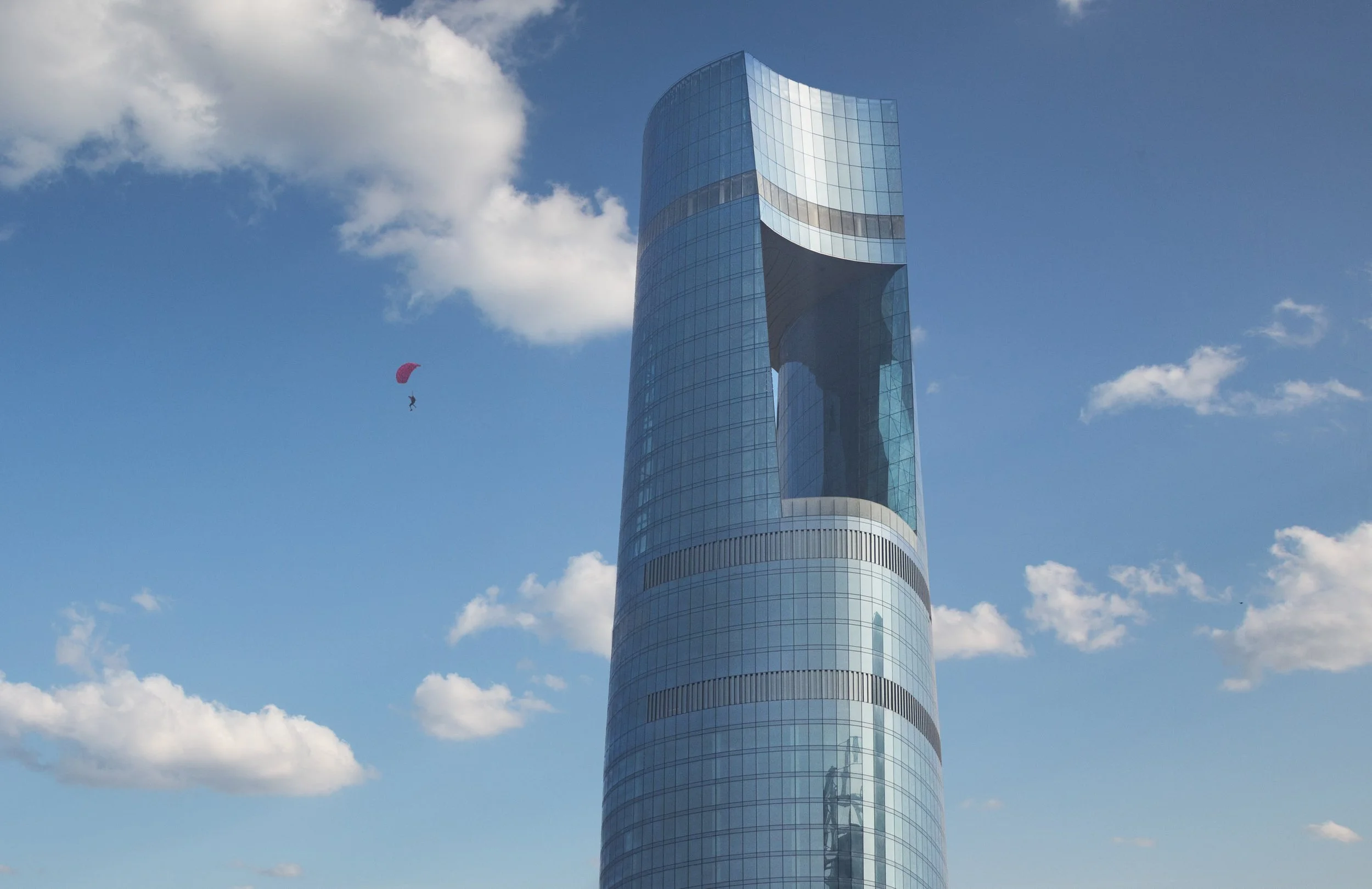 The crown and aperture of Ciel Dubai Marina against cloud, a parachute crossing the sky at left.
