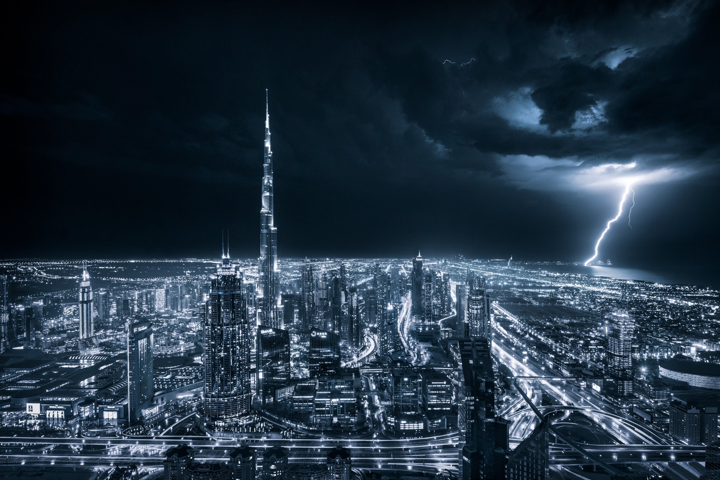 Nighttime cityscape of Dubai with illuminated skyscrapers, including the Burj Khalifa, under a stormy sky with lightning - fine art photograph by Ahmad Alnaji, SARAB Art Production