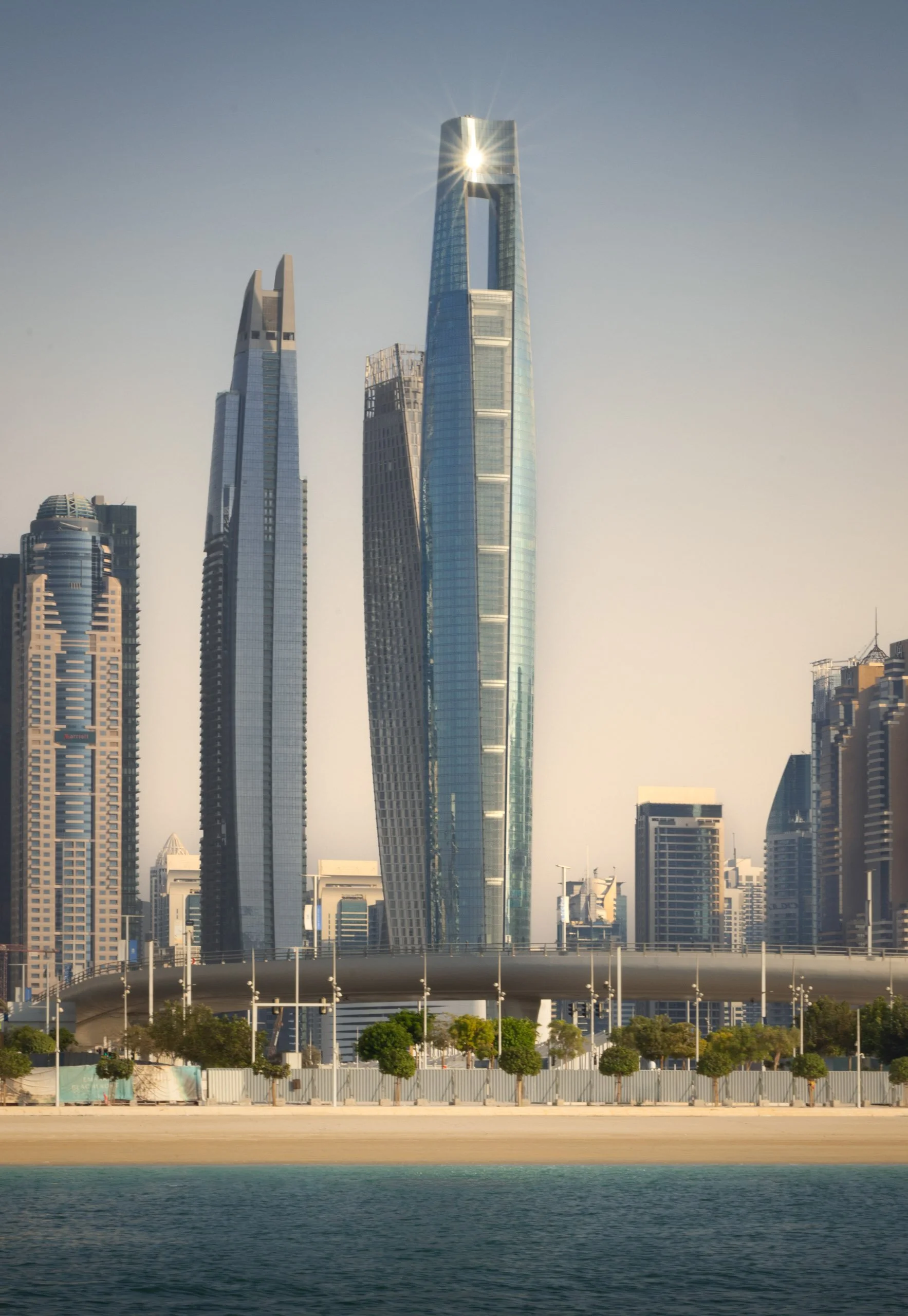 The sun flaring at the crown of Ciel Dubai Marina, the bridge and the shoreline below.