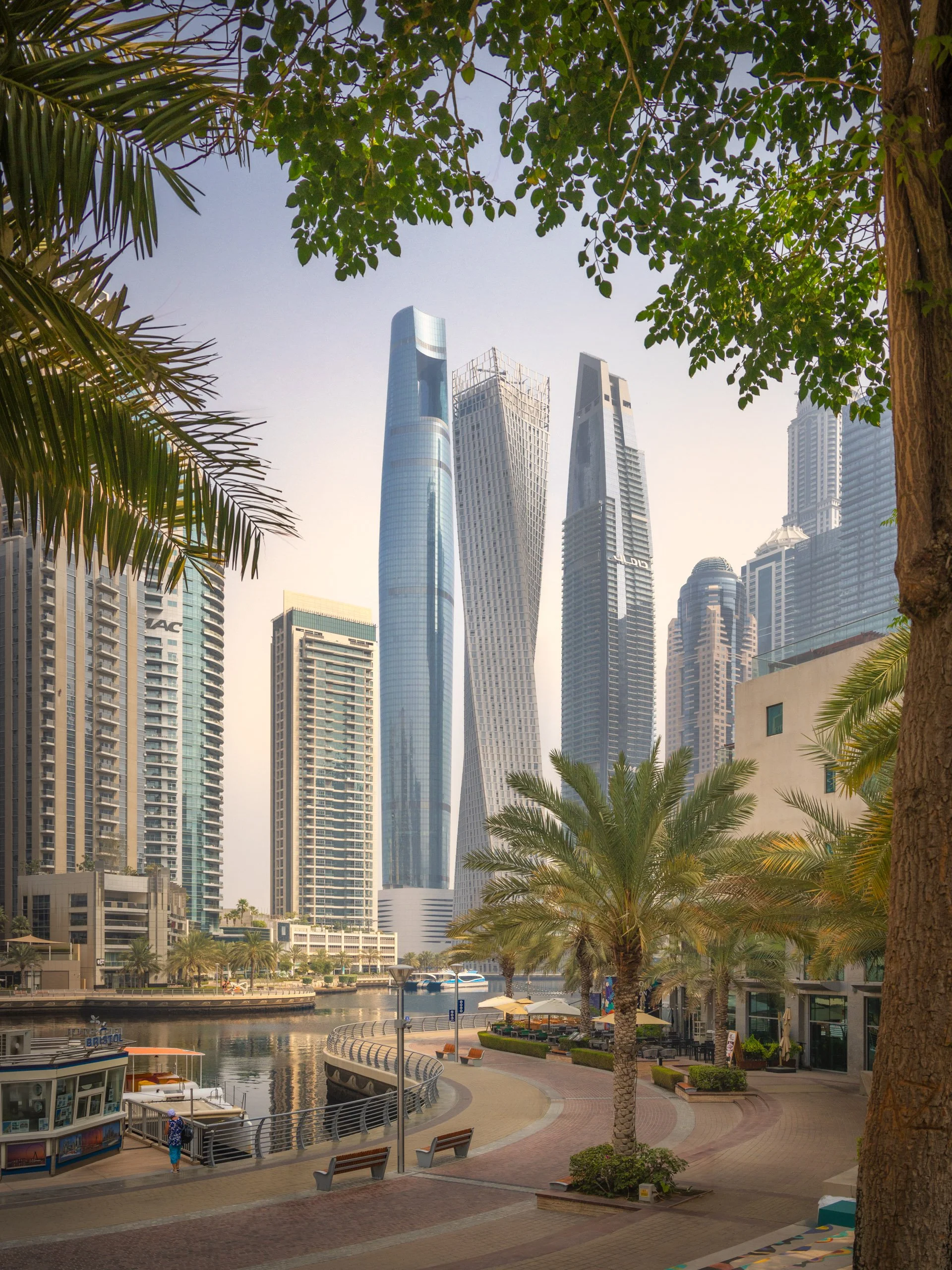 Ciel Dubai Marina framed by trees along the marina walk, boats moored on the water below.