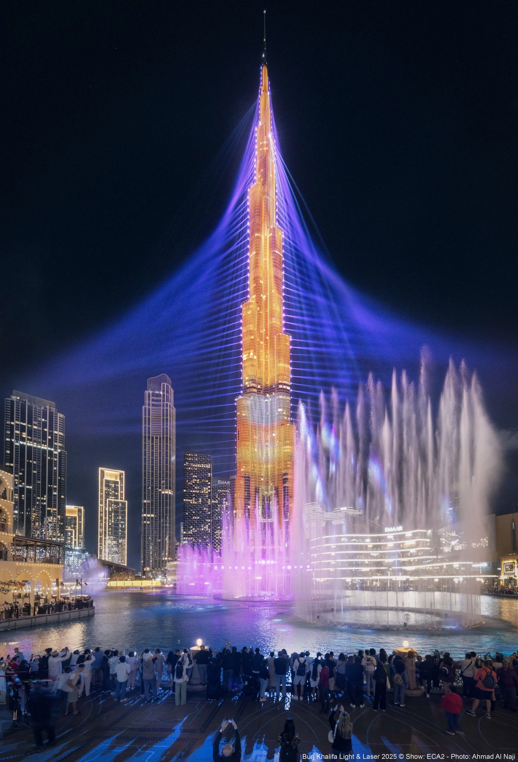 Nighttime view of the Burj Khalifa in Dubai illuminated with its colorful light show, with a fountain display in the foreground and a crowd of people watching - fine art photograph by Ahmad Alnaji, SARAB.