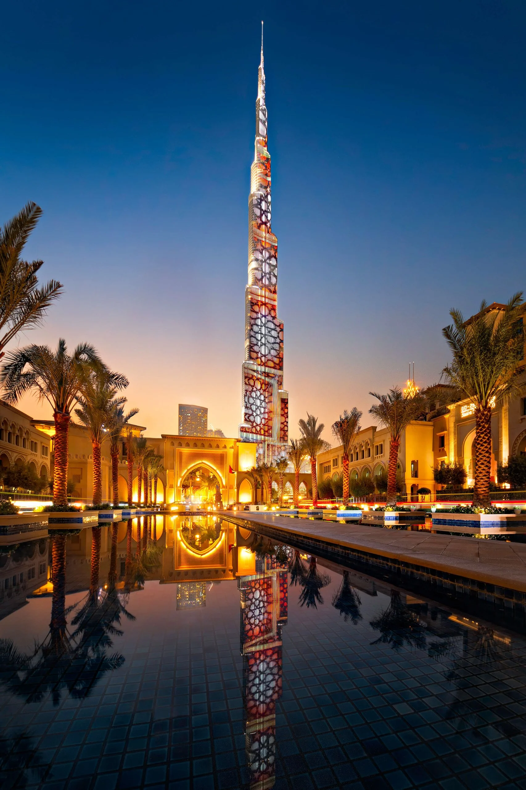 Night view of the Burj Khalifa with colorful lighting, surrounded by palm trees and historic buildings, reflected in a water pool.