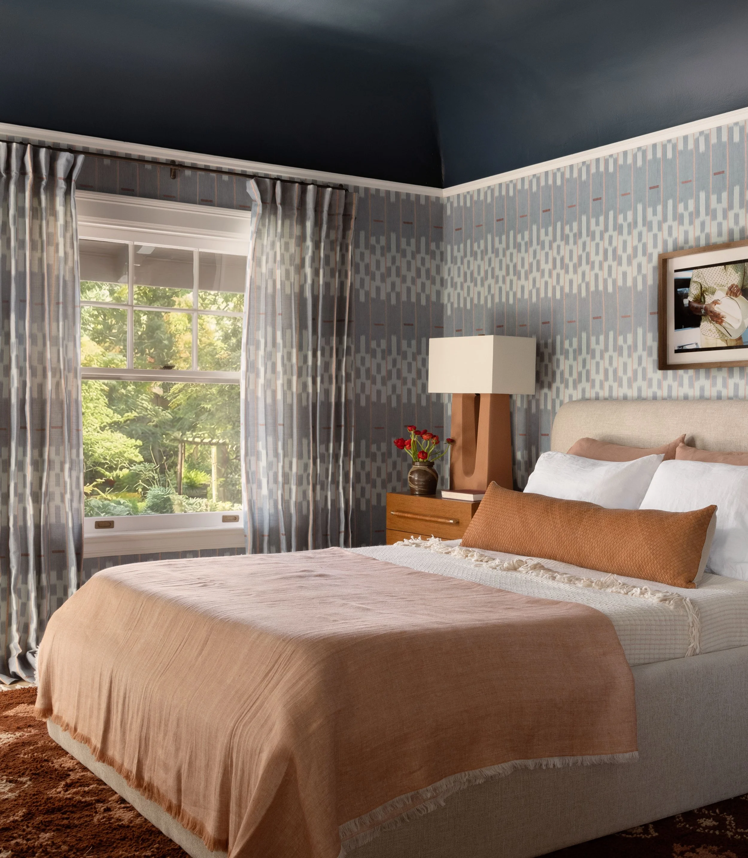 Close-up of a bedroom bedside table with a vase of red flowers, a lit candle, and a stack of books. Part of the bed with a decorative pillow and a textured throw blanket is visible.