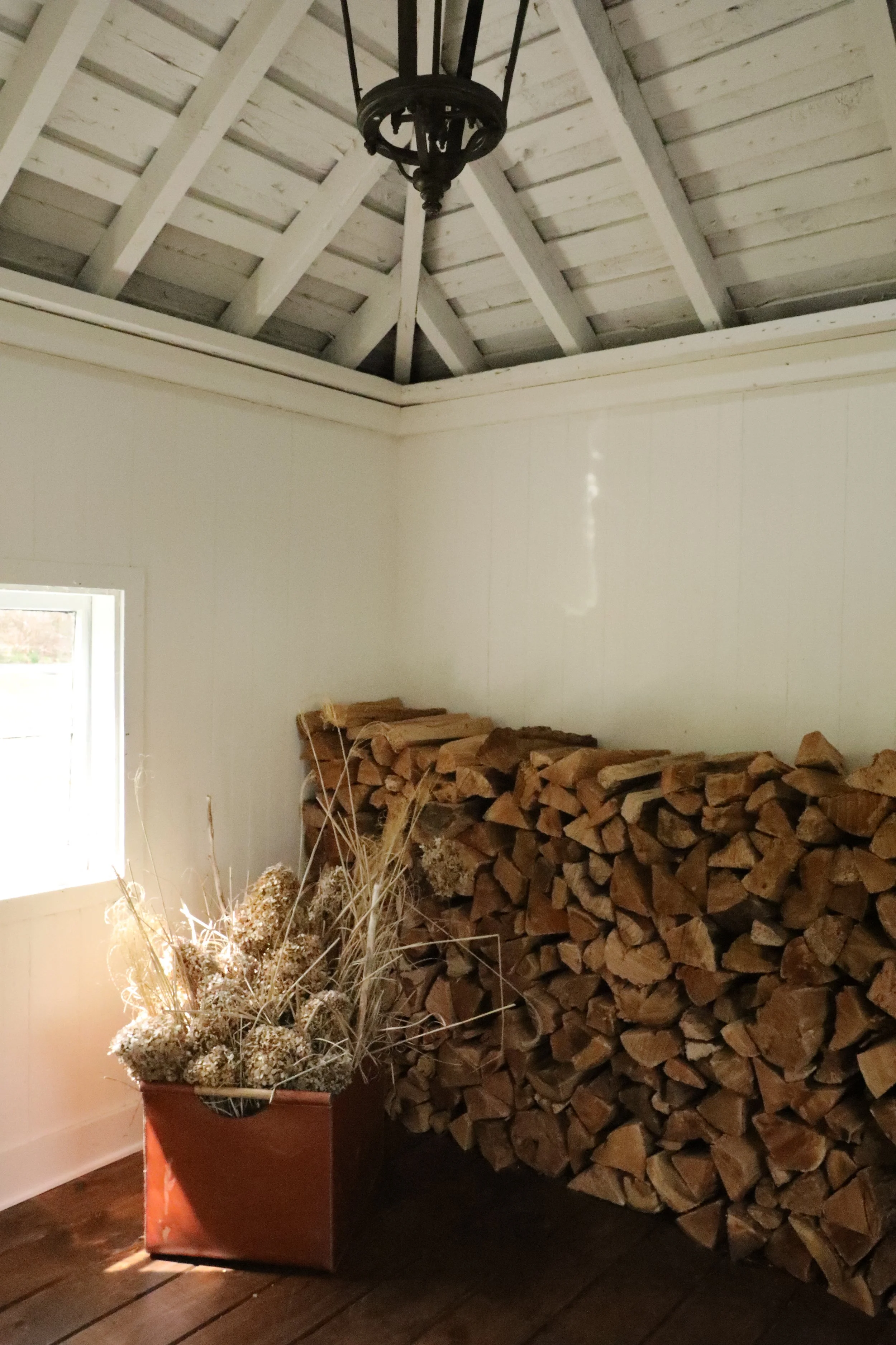 Interior of a room with a large stack of firewood, a potted dried flower arrangement, white walls, wooden floor, and a chandelier hanging from the ceiling.