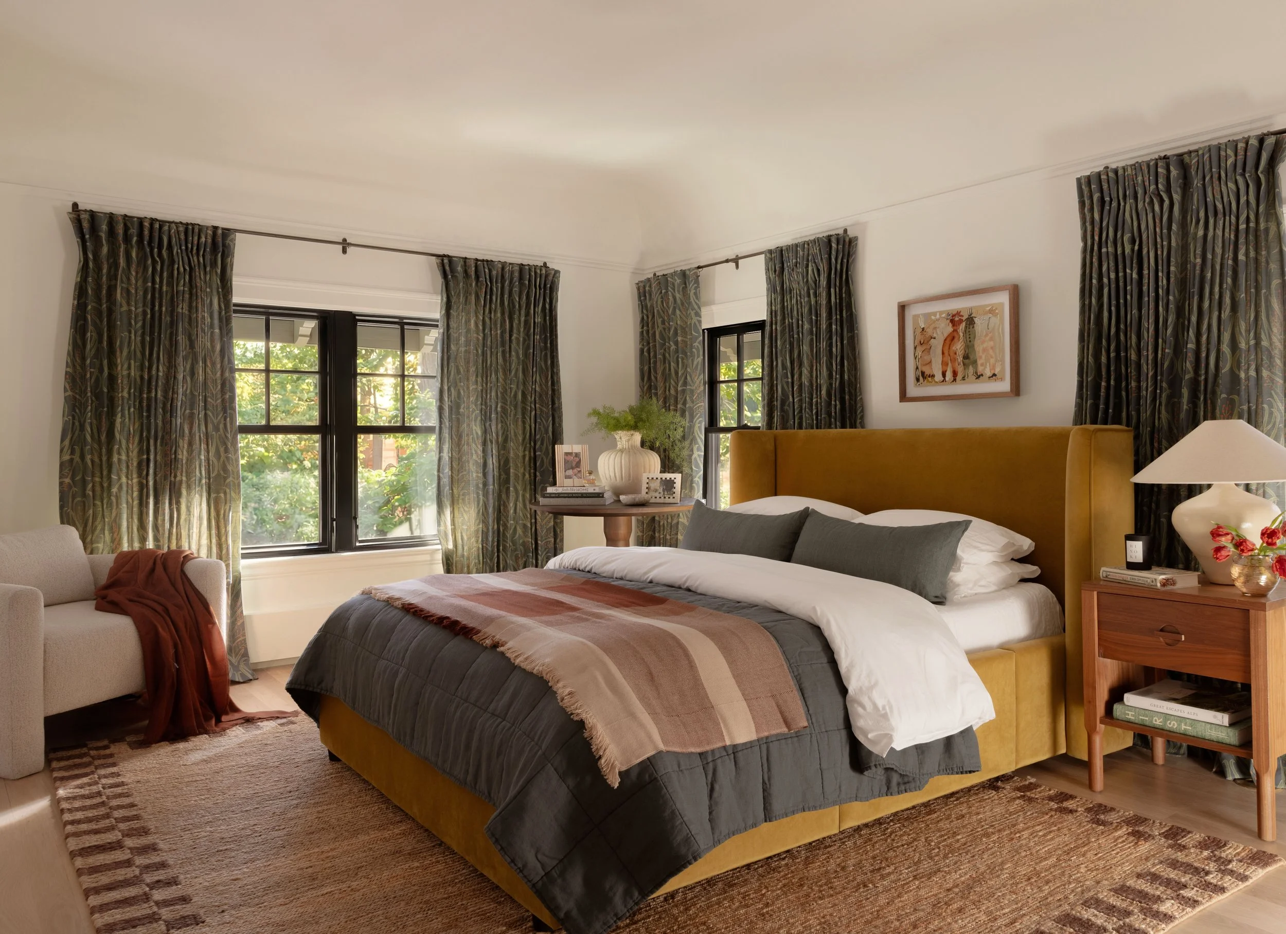 A cozy bedroom featuring a bed with a mustard-colored headboard, dark gray bedding, and a striped blanket. There is a wooden nightstand with a lamp, flowers, and books, and a window with green, patterned curtains. A beige armchair and a woven rug are